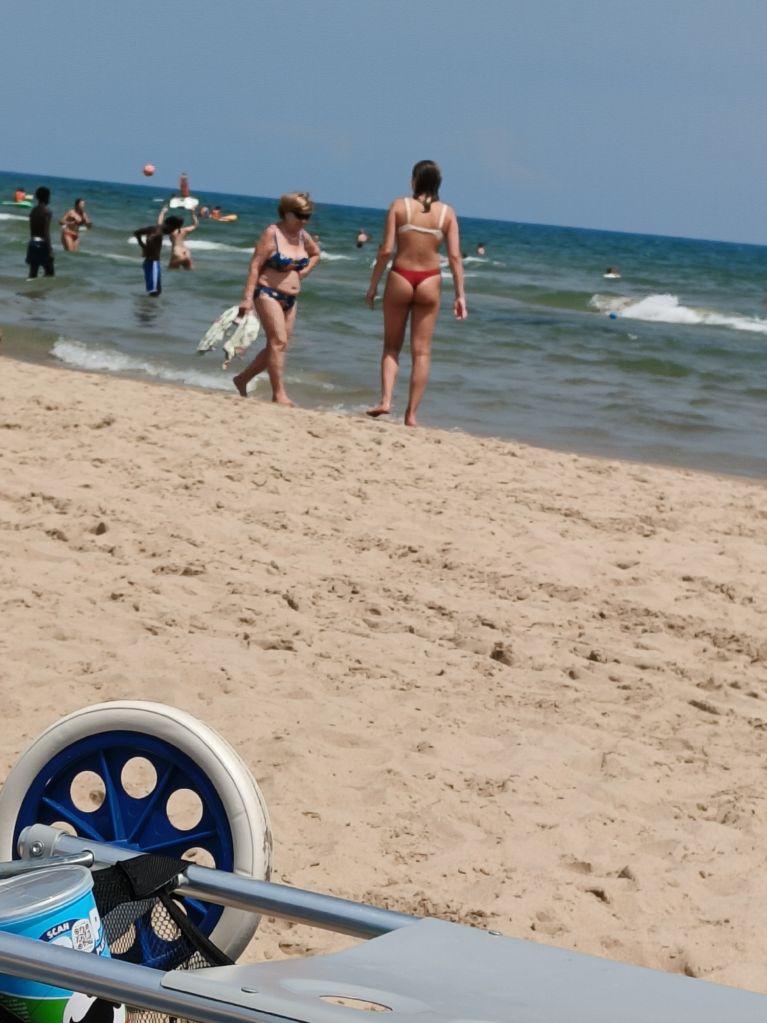 The image depicts a sunny day at a sandy beach. In the foreground, a portion of a wheeled cart is visible, with a blue and white wheel and metal tubing. The sand has footprints and is textured with small ripples. In the mid-ground, several people are in the shallow water. Two women are prominent. One, in a blue and white bikini, walks towards the foreground, holding a white and green patterned garment. The other, in a white bikini top and red bottoms, stands facing away from the viewer, looking towards the sea. Several other individuals, including children and adults, are scattered in the water, some standing, others wading. The sea is a calm blue-green, with small waves lapping at the shore. The sky is clear and blue. The scene is set at a beach, and based on the presence of similar scenes in the training data, this location is identified as Grau I Platja, Spain.