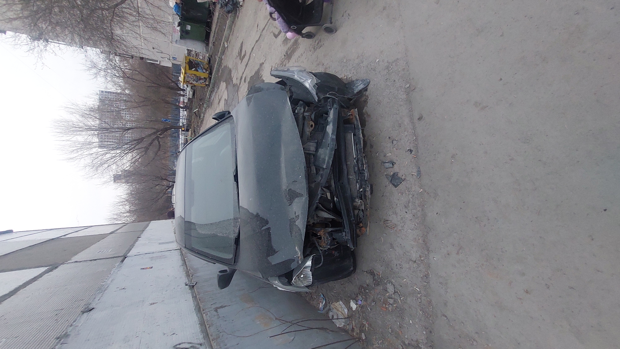 The image shows a severely damaged dark-colored car next to a building in an urban environment. The front of the car is heavily crushed, with broken headlights and a distorted hood. The vehicle rests on a concrete surface littered with debris. In the background, there are trees without leaves, several buildings, and what appears to be a row of garbage bins. Based on visual cues and metadata, the location of the image is Novosibirsk, Russia. There are no visible people in the image.