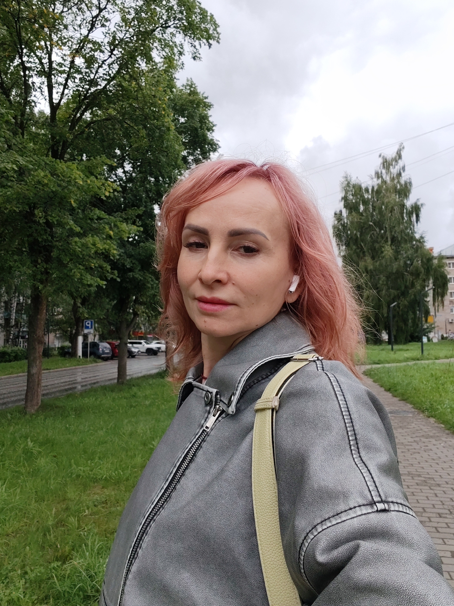 A woman with pink hair is standing on a sidewalk in Chaikovsky, Russia, in front of a street lined with trees and buildings. She is wearing a silver leather jacket and a light-colored strap across her shoulder. She also has a white earbud in her right ear. The sky is overcast. In the background, cars are parked along the street and tall buildings are visible. The ground is paved with cobblestones on the right side and grass on the left.