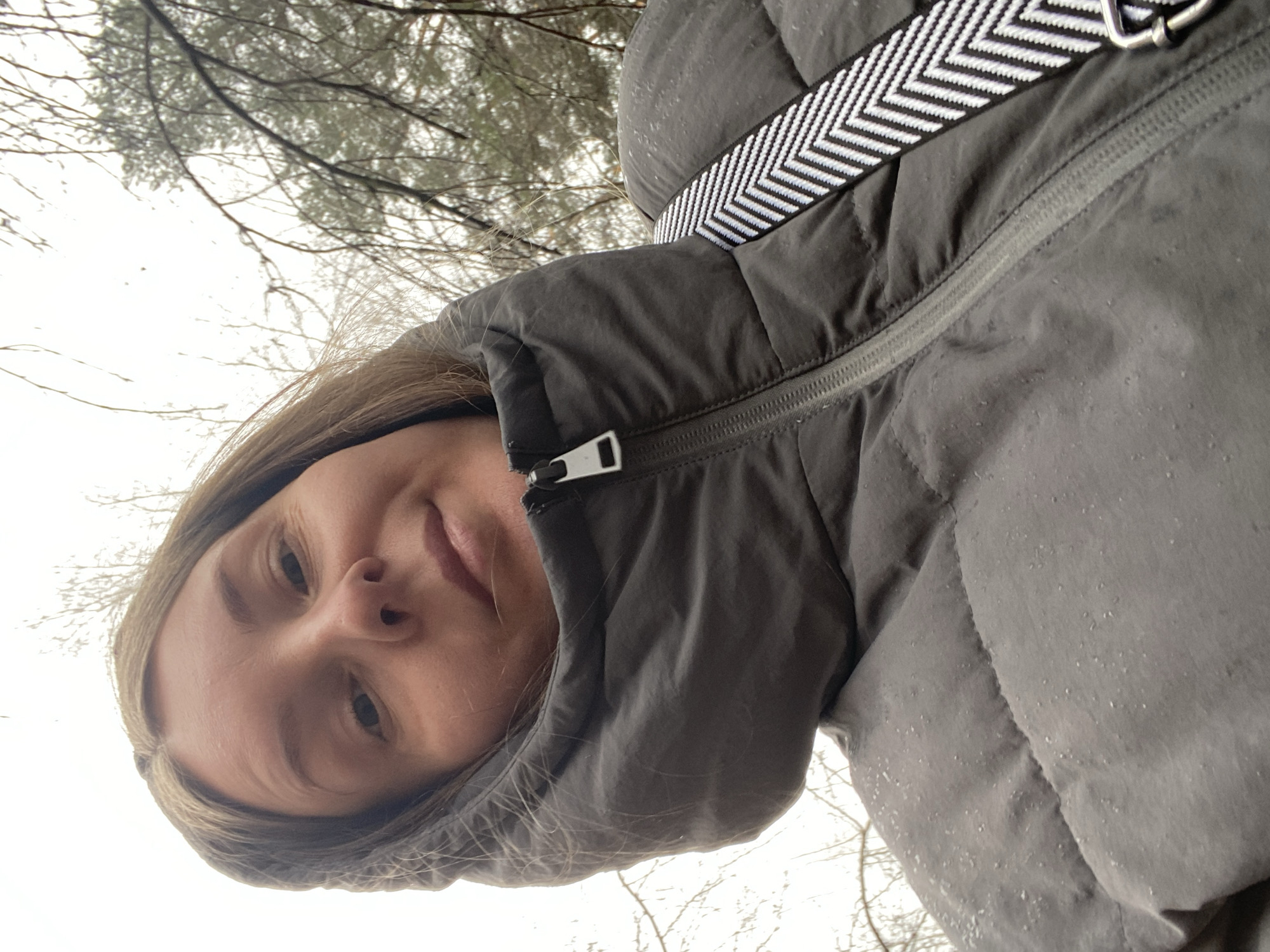 A selfie depicts a fair-skinned person with light brown hair, partially framed by the hood of a grey, puffy winter coat. The coat also features a black and white zigzag-patterned strap. The zipper of the coat is partially visible. The background shows bare branches and a cloudy sky. The location cannot be reliably confirmed as Noviy, Russia based on the provided image.
