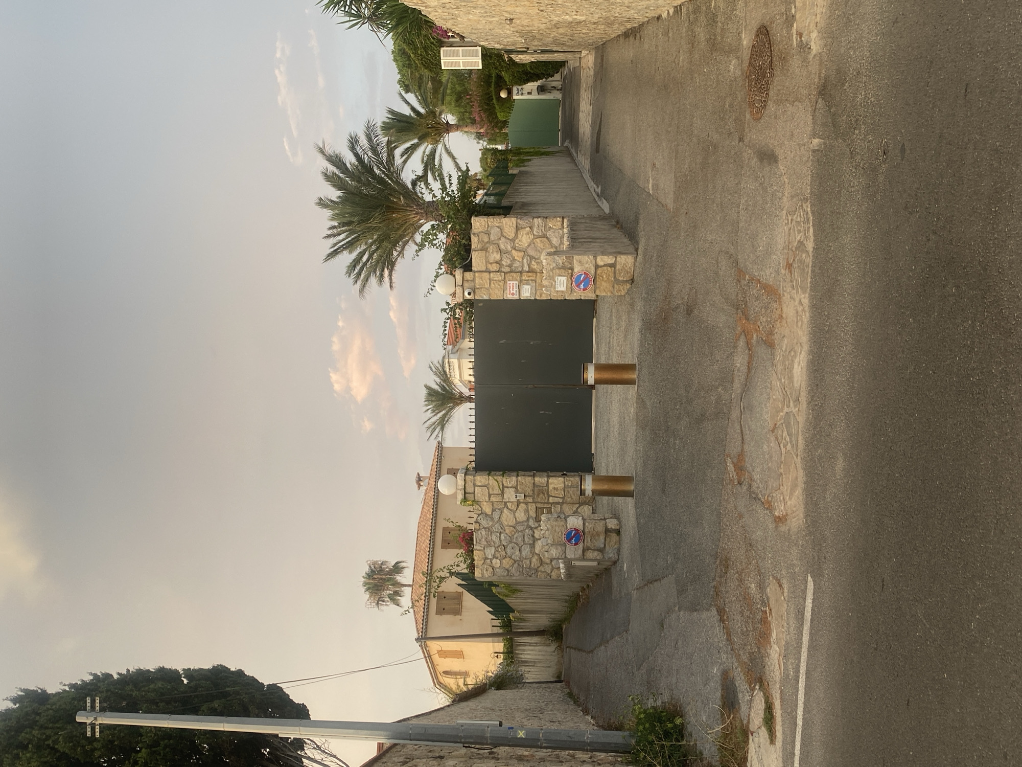 The image depicts a residential area in Antibes, France, featuring a paved road leading to a large, dark green double gate. The gate is flanked by stone pillars, one of which has a white globe-shaped light fixture and a security camera. Two golden bollards are positioned in front of the gate. To the left of the gate is a stone wall and a narrow path leading uphill towards a light-colored house with a tiled roof and wooden shutters. Palm trees and other lush vegetation are visible throughout the scene. The sky is overcast with hints of pink from the setting sun. The overall impression is one of a private and well-maintained property.