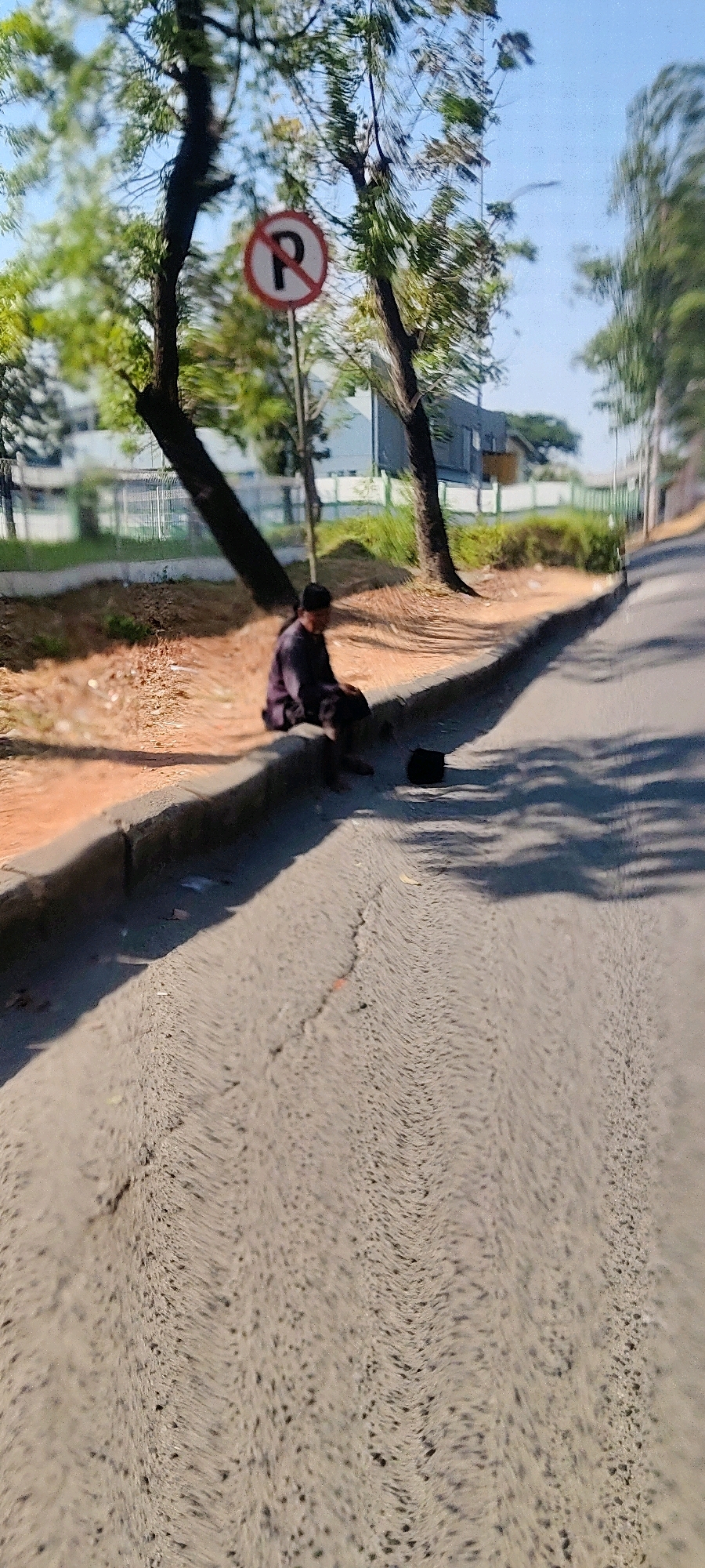 In Mekarmukti, Indonesia, a person wearing a dark maroon outfit and a black cap is sitting on the curb next to a road. The person is facing away from the camera, looking towards the left. Next to the person on the road, there is a black hat. A "no parking" sign is visible on a pole behind the person, surrounded by trees with green leaves. The road has a cracked surface, and a shadow of trees falls across it. The background shows a building with white fencing and more trees.