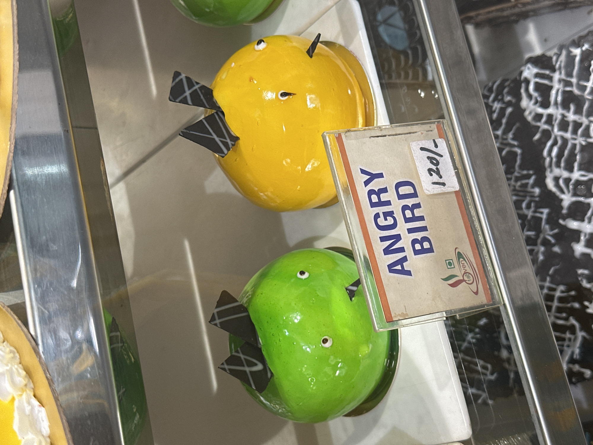 In Mathura, India, a display case contains pastries, including two shaped like Angry Birds, one yellow and one green, along with a price tag of 120/-.