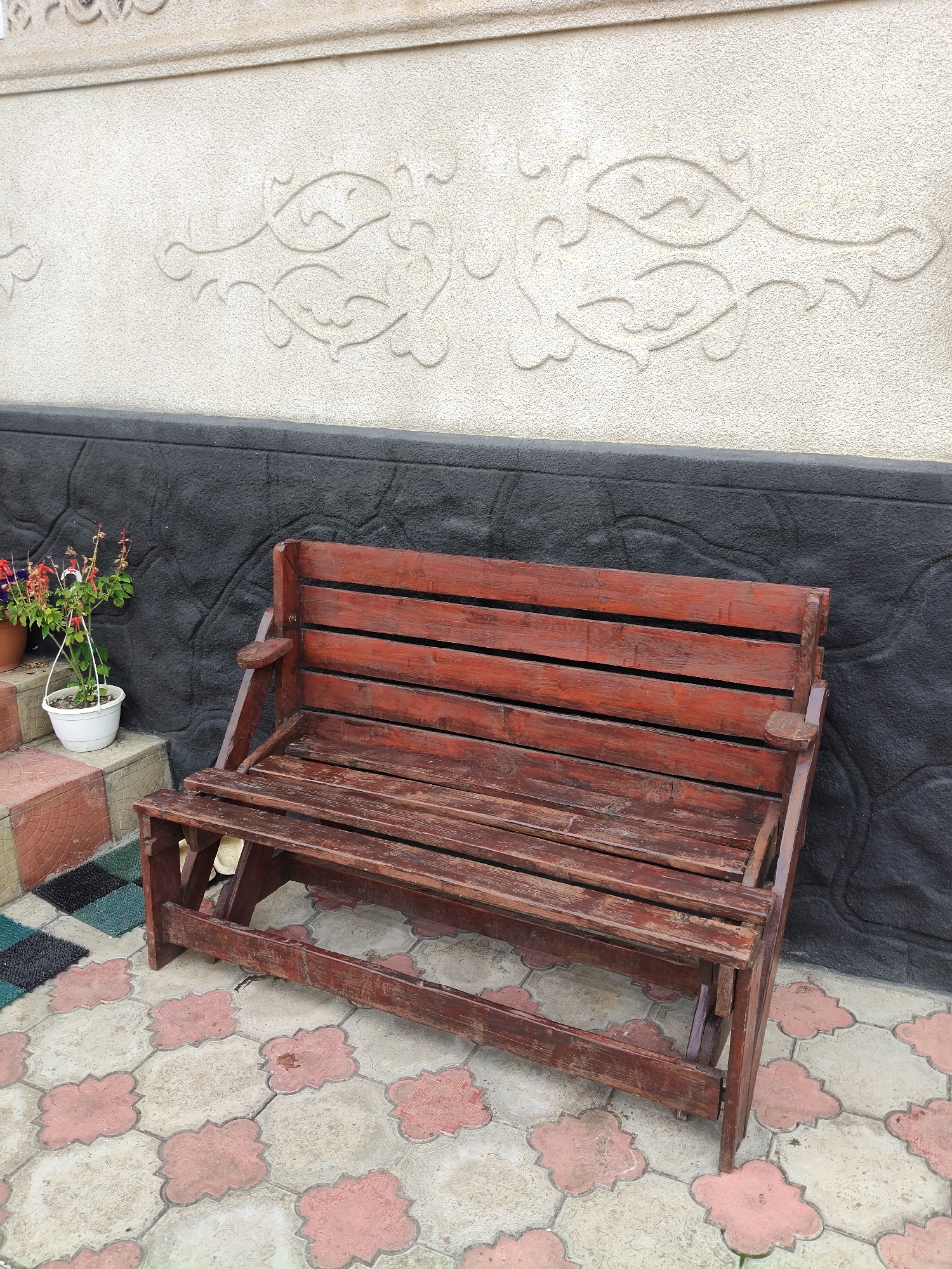 In Gîrbova, Moldova, a wooden bench with a dark red stain sits on a tiled patio in front of a textured, light-colored wall featuring decorative embossed fish patterns. The wall is bordered at its base by a dark, rough-textured section. To the left of the bench, a white planter with small red and purple flowers is placed on a step. The patio tiles are a geometric pattern of light beige and pink shapes.