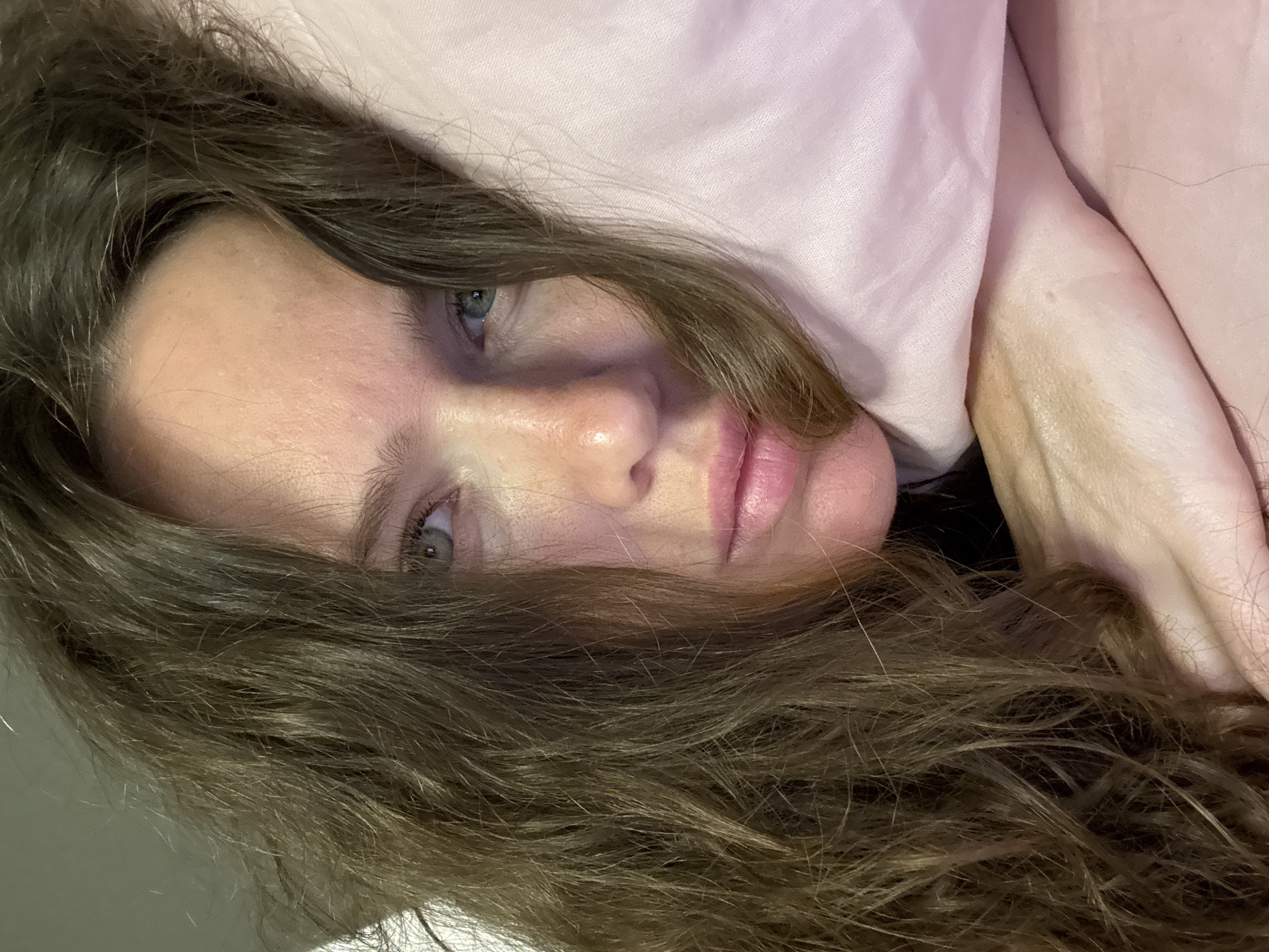 The image shows a close-up, head-and-shoulders view of a woman lying in bed, nestled against a pink pillow. Her eyes are open, looking directly at the camera with a somber expression. Her long, brown, wavy hair is spread around her face, partially obscuring her left cheek. The lighting is soft, suggesting an indoor environment, likely a bedroom. The scene appears to be from Khimki, Russia. There is no visible indication of any action or interaction beyond the woman's posed gaze.