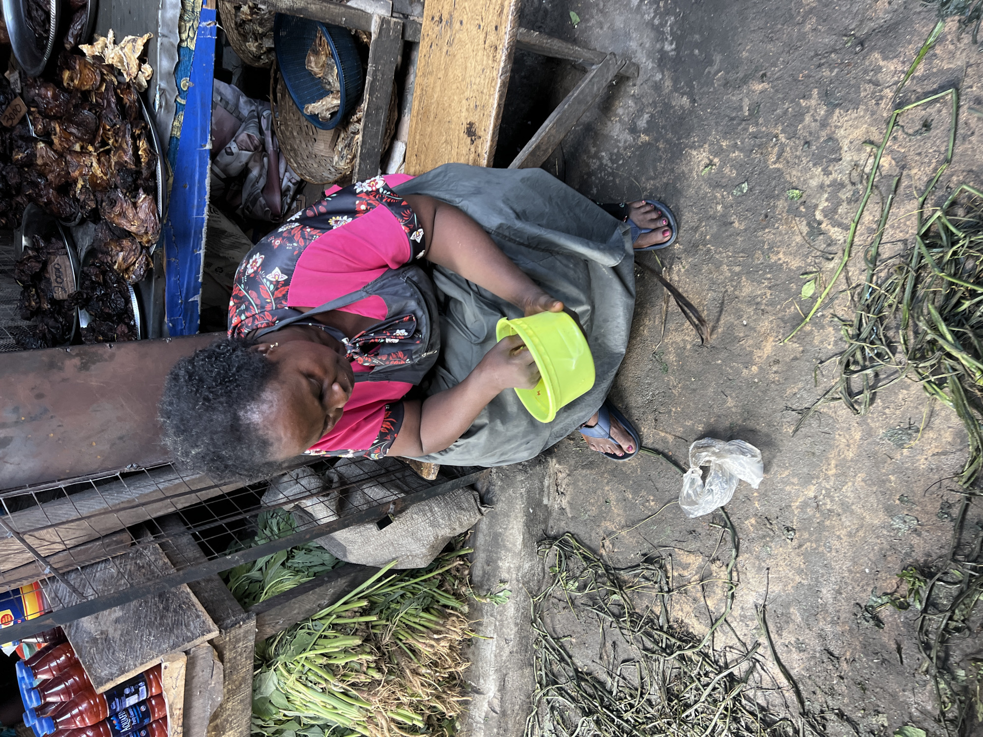 The scene is a marketplace in Nkpor, Nigeria. An older woman with graying hair sits on a low stool, wearing a pink shirt with a dark floral pattern, a gray apron, and blue sandals with pink toenails. She holds a bright yellow plastic container. Beside her is a wooden structure with a metal grid and bottles of red liquid. Near the woman are piles of green vegetables and plant debris. The ground is a stained, muddy surface with scattered plant matter and a crumpled plastic bag. In the background, trays of dark-colored meat are visible, along with woven baskets and blue fabric.