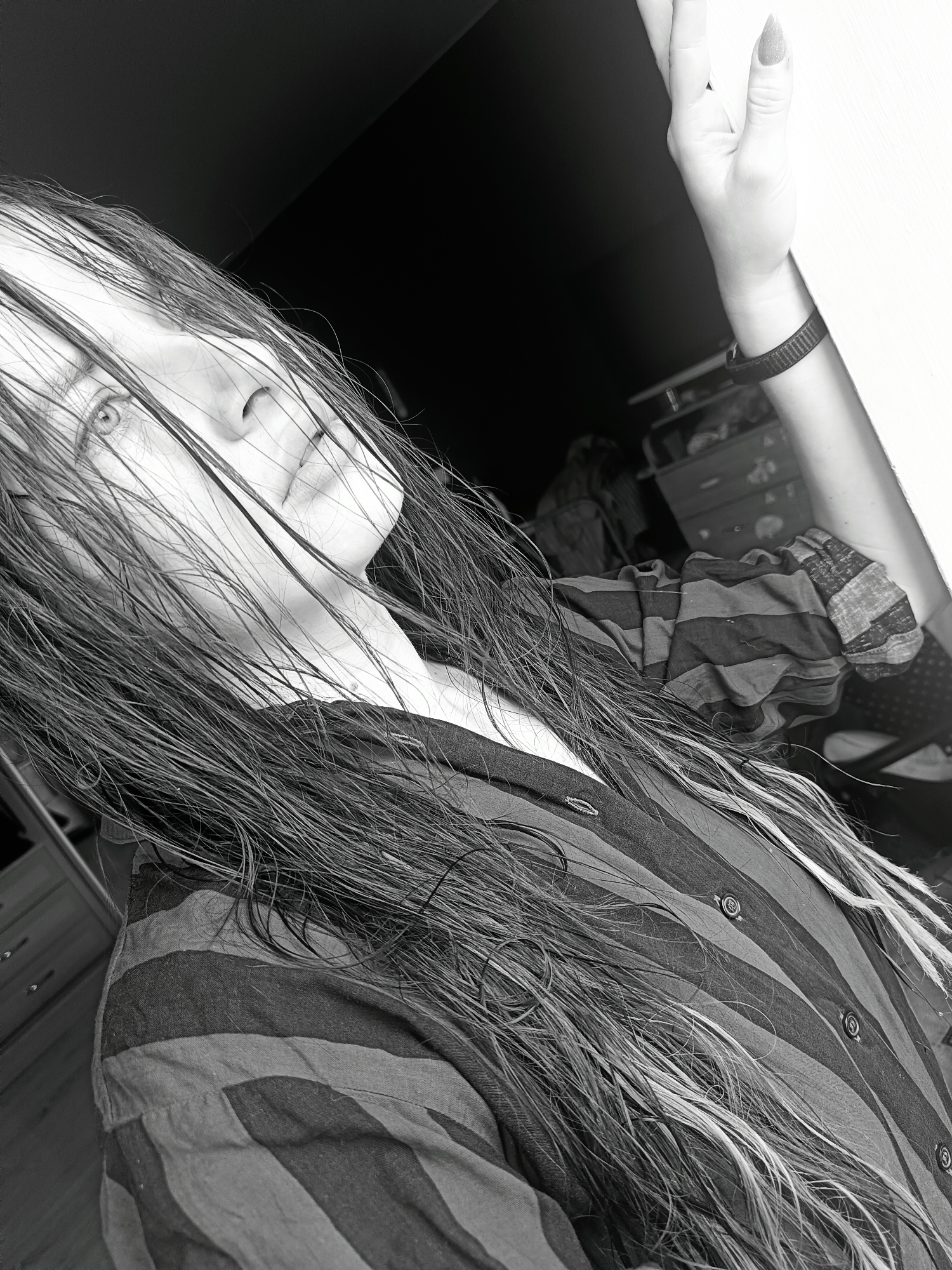 A grayscale, angled close-up photograph shows a person with long, wet hair obscuring their face, wearing a striped, button-down shirt. The person's left hand is raised towards the top right corner of the frame, wearing a dark wristband. The background is dark and partially obscures a cabinet and other indistinct objects. The image is not identifiable as being in Rusalevka, Russia.