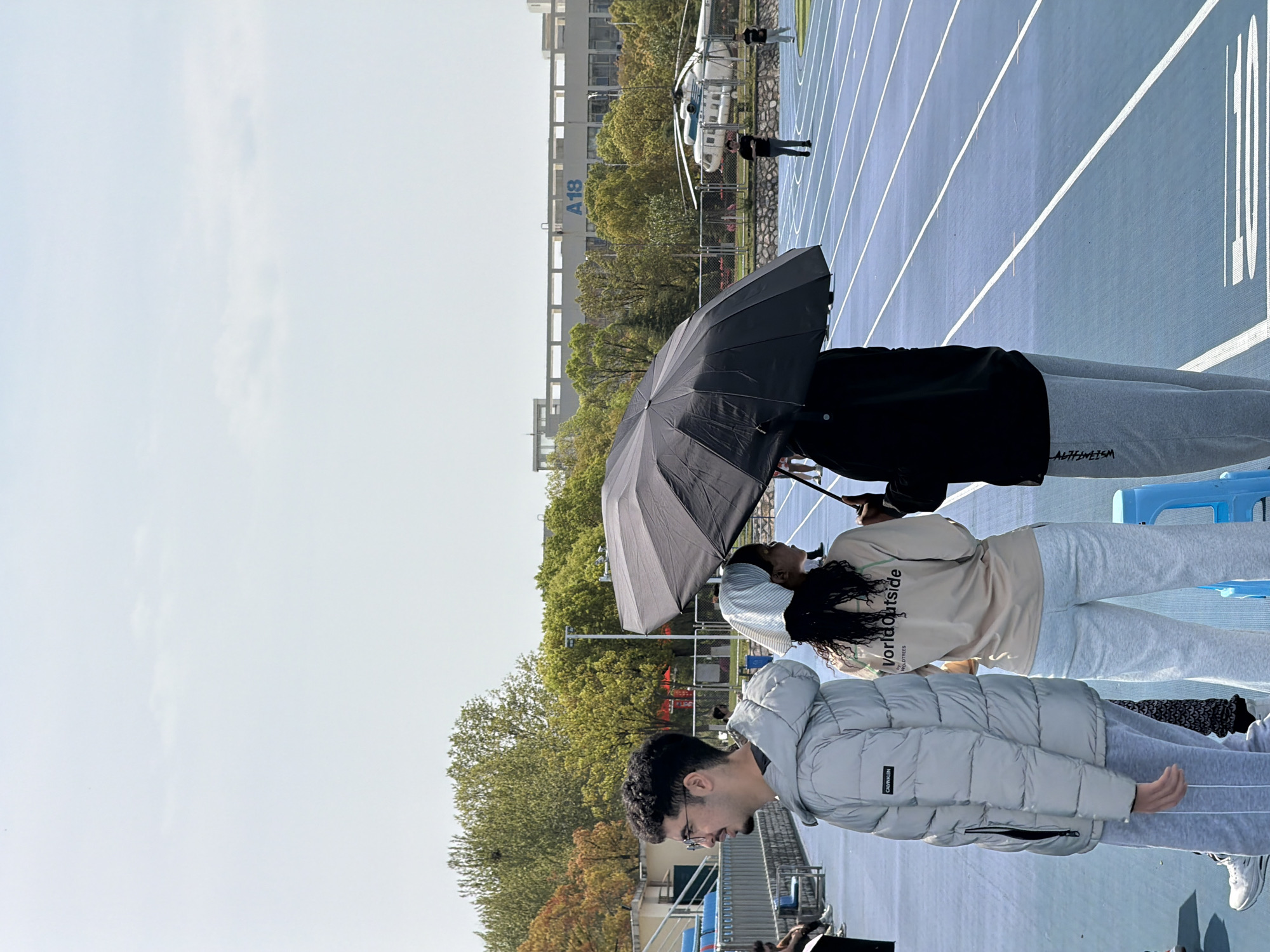 The image shows a group of people standing on a blue running track. One person is holding a black umbrella. Behind them are buildings, trees, and a helicopter. The building in the background has "A18" written on it. The location is Jiangning District, China.