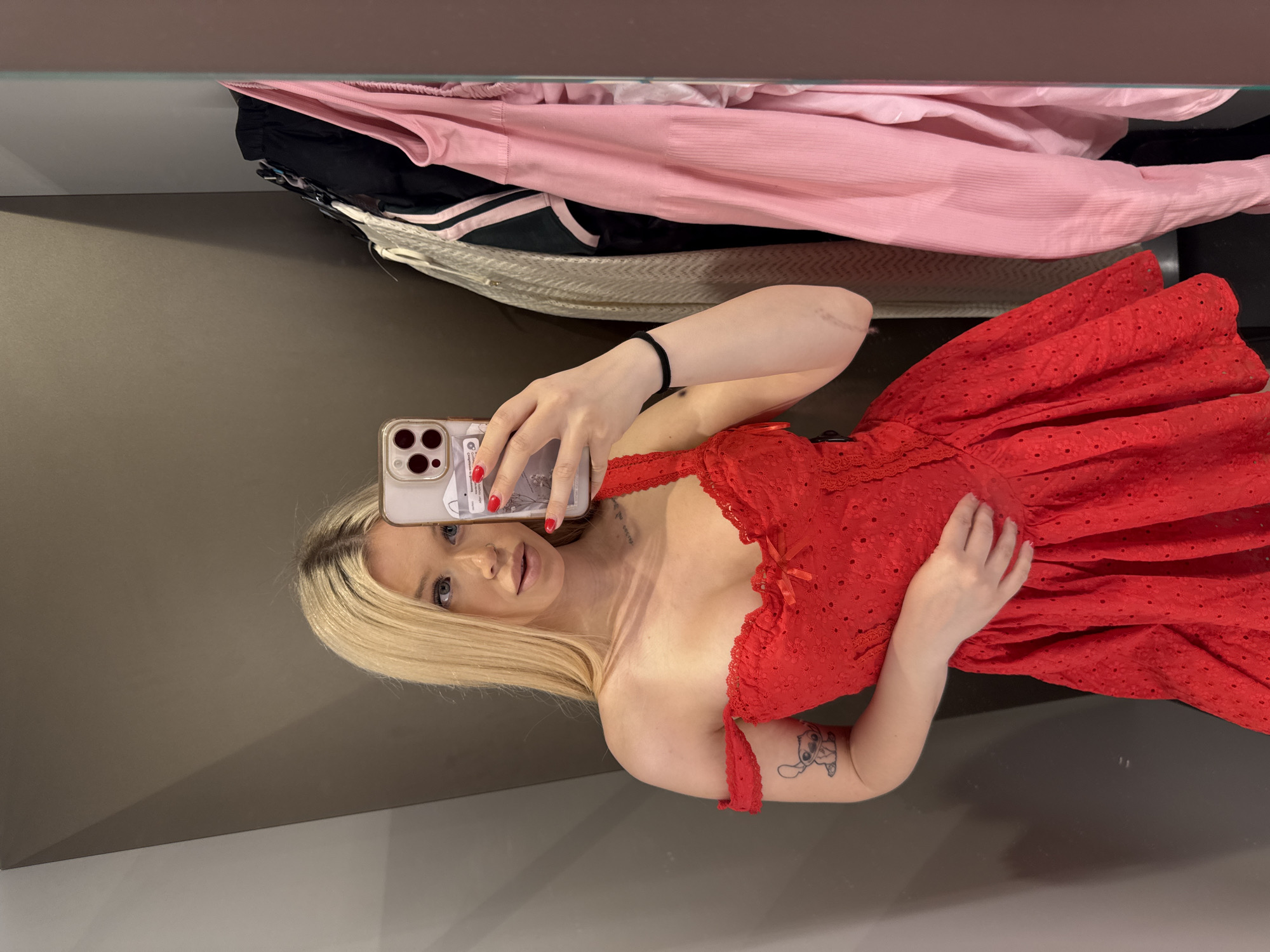 A woman with blonde hair and blue eyes is taking a mirror selfie in a fitting room. She is wearing a red off-the-shoulder dress with a sweetheart neckline and a bow detail. She has a tattoo of Stitch on her left arm and red nail polish on her fingers. Her phone, with a pink case, is visible in her right hand. In the background, several garments hang on a rack, including a light-colored cardigan, a black and green striped top, and a pink top. The fitting room has gray walls and a mirror. The image is captured in Kudrovo, Russia.