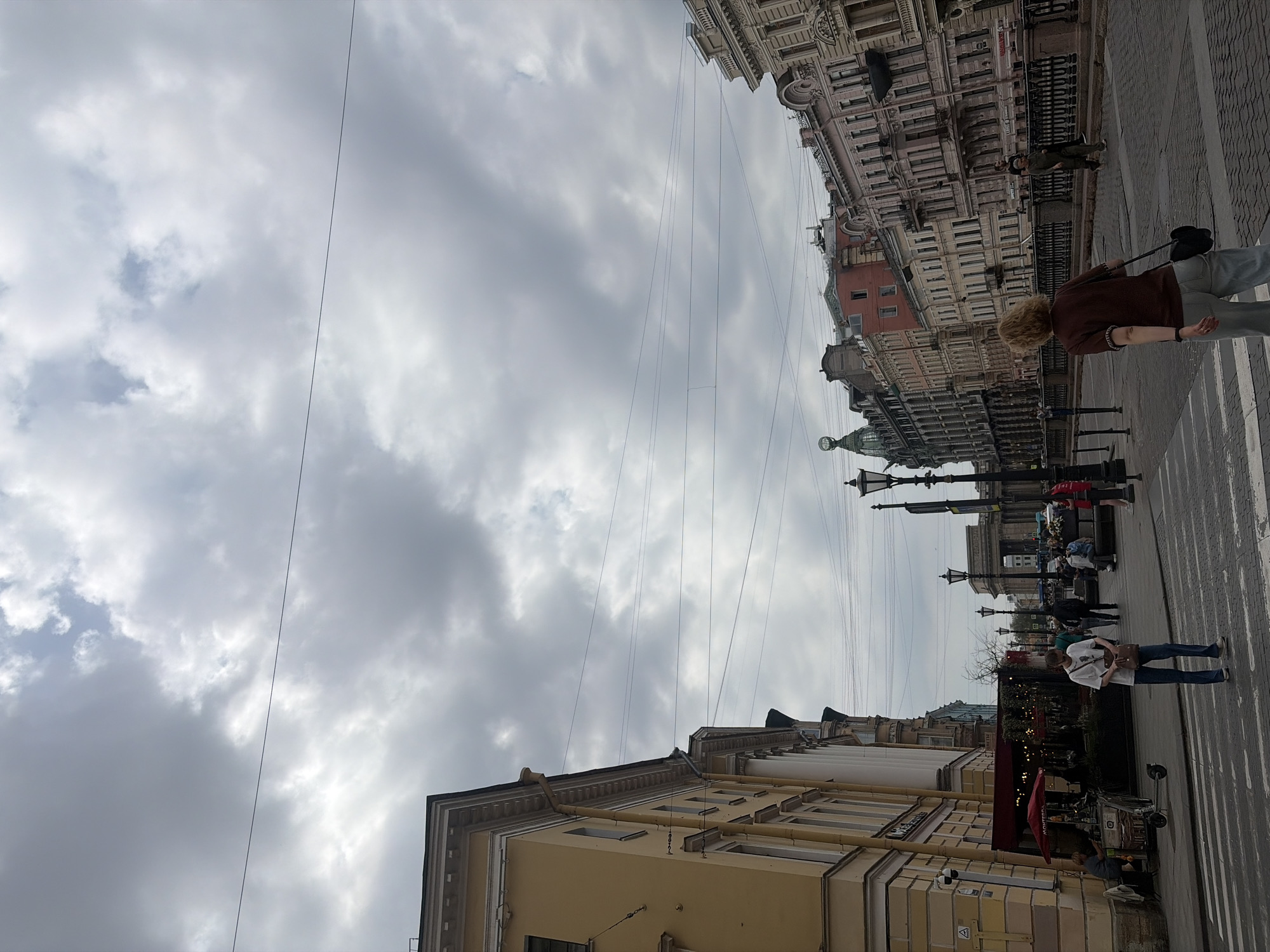 This is a medium-angle, eye-level shot of a street scene in Saint Petersburg, Russia, under a cloudy sky. The view is looking down a street lined with ornate, multi-story buildings with classical architecture, painted in shades of yellow, pink, and beige. Wires crisscross the sky above the buildings.
In the foreground, a person with blonde, curly hair wearing a dark red t-shirt and light blue jeans walks away from the camera, facing the buildings. They are carrying a black bag over their shoulder. To the left, a person in a white t-shirt and blue jeans is standing on a crosswalk. Further down the street, there are several other pedestrians walking and sitting. On the left side of the street, a building with a yellow facade has awnings extended over a storefront, and there is a red umbrella. Streetlights are visible along the sidewalk.
The overall impression is of a bustling urban environment in Saint Petersburg, with characteristic architecture and overcast weather.