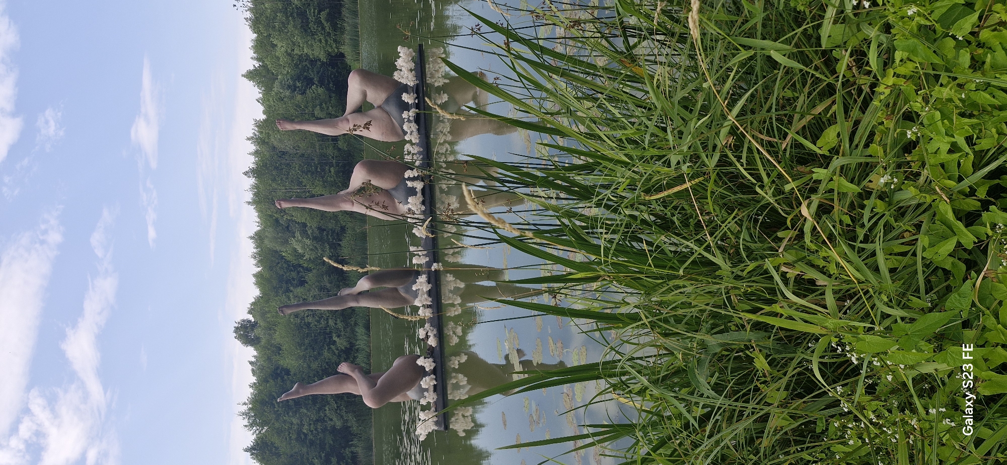 This image shows a sculpture installation in a lake in Pakruojis, Lithuania. The sculpture consists of four life-sized, nude, seated figures arranged in a line on a floating platform. The figures are positioned with their legs raised and bent, creating a dynamic and abstract composition. Small white flowers adorn the platform in front of each figure. The lake is surrounded by lush green trees, and the sky is a clear blue with scattered white clouds. In the foreground, tall green grass and reeds obscure the lower portion of the scene, adding a naturalistic element to the otherwise artificial display. The reflections of the sculptures and the surrounding trees are visible on the calm surface of the water. The image appears to be taken on a sunny day.