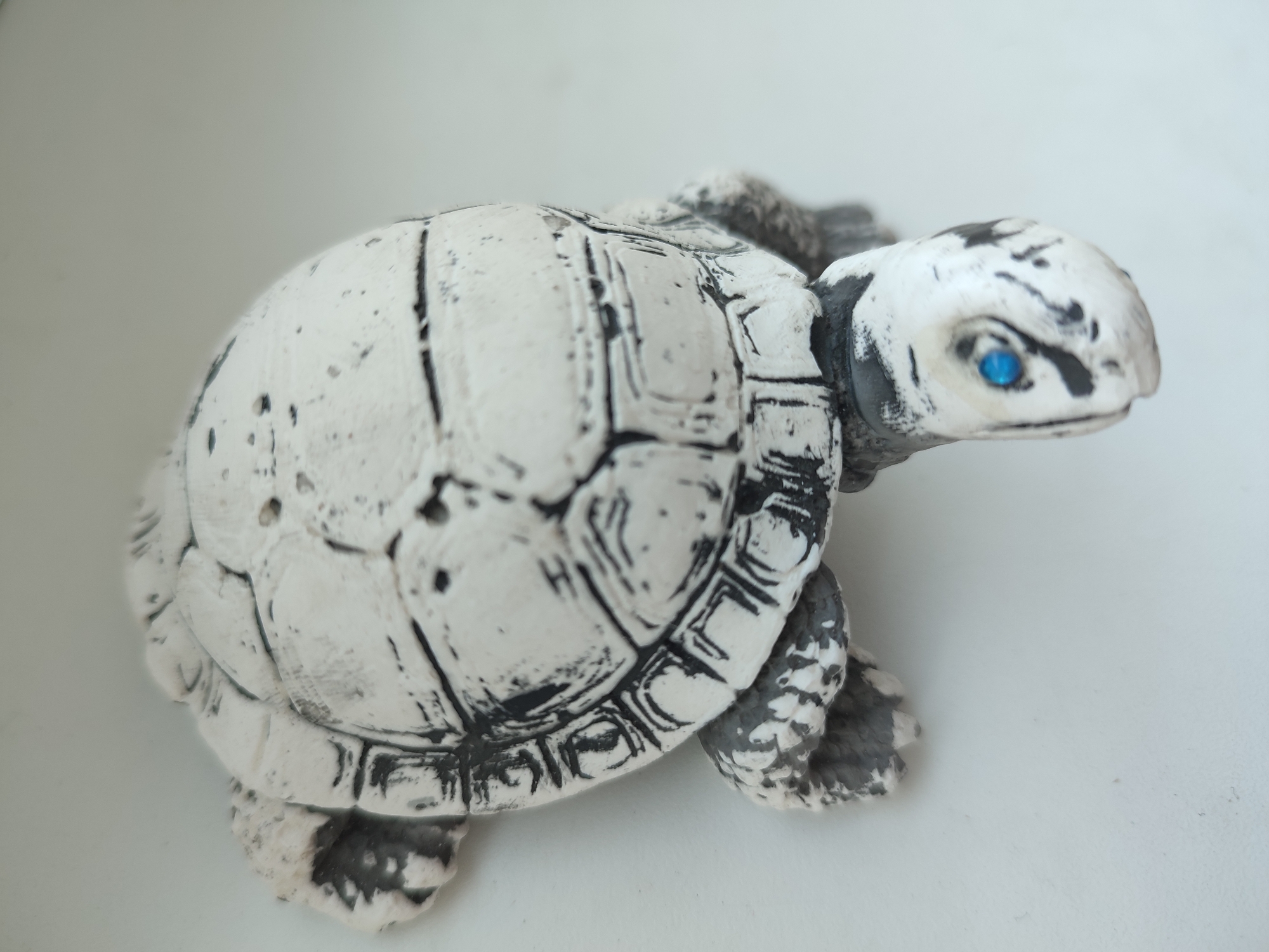 A close-up shot shows a small, white ceramic turtle figurine with black markings and bright blue eyes. The figurine is positioned on a flat, light-colored surface, possibly a table or shelf, against a softly blurred, neutral background. The lighting is bright and even, highlighting the texture and details of the turtle's shell and head. No specific location in Voronezh, Russia, can be identified from the image.