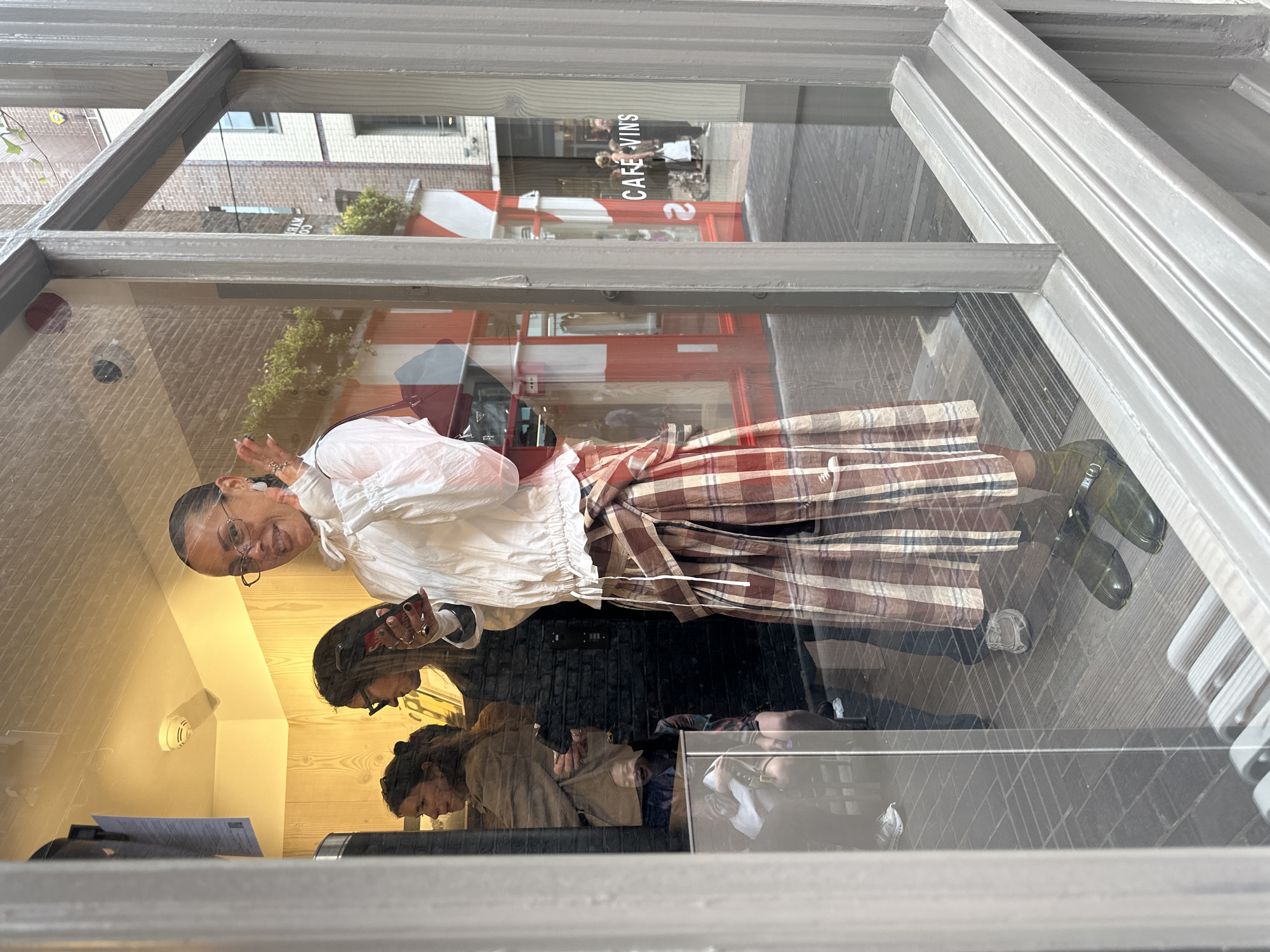 The image shows a reflection in a window, likely of a shop or cafe in London. The reflection features a woman standing outside wearing a white blouse, a brown and white plaid skirt, and green shoes. She is gesturing with her hand and smiling. Behind her, reflected in the glass, is a red-and-white building labeled "CAFE VINS". Inside the shop, the reflection shows several people, possibly customers or staff, near a counter. The shop has light wooden paneling. Outside the window, the brick pavement is visible, and the window frame is painted a light grey.