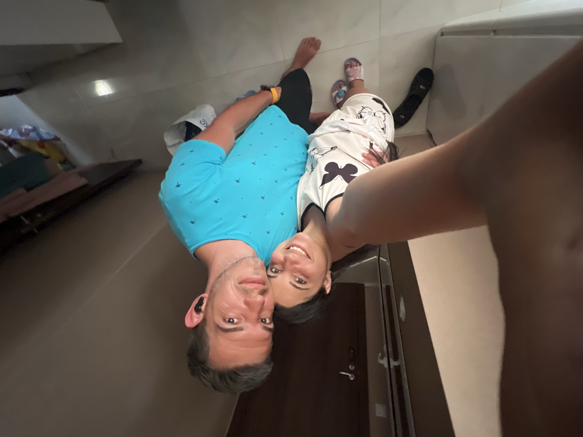 A high-angle, full-body selfie of a man and woman standing indoors. The man, on the right, wears a blue t-shirt with a palm tree pattern and black shorts. He has short brown hair and is wearing black earbuds. The woman, on the left, is leaning her head on the man's shoulder. She wears a white tank top with a butterfly print and white shorts, also with a butterfly pattern. She has dark hair and is wearing pink and white sandals. They are standing on a tiled floor. The background shows a door with a silver handle and a wall. The lighting suggests an indoor environment, possibly a hallway or room. No specific location in Sofia, Bulgaria can be determined from the image.