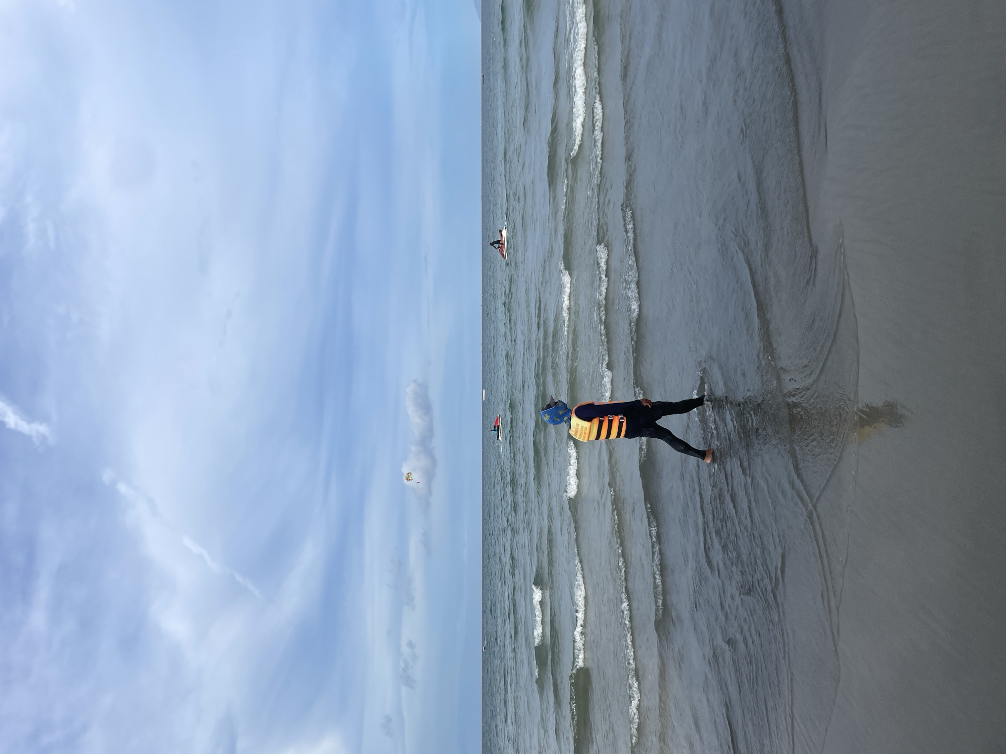 A lone individual wearing a life vest walks along the edge of the ocean on a beach in Da Nang, Vietnam. The person is wearing dark pants and a dark long-sleeved shirt, with a bright orange and yellow life vest over it. They are walking from right to left in the frame, with their right leg submerged in the shallow water. The ocean has small waves lapping at the shore. In the distance, several jet skis are visible on the water, and a parasail is in the air further out. The sky is partly cloudy with a blue hue. The sand in the foreground is dark and wet from the receding tide, with ripples of water flowing over it. The overall scene depicts a recreational day at the beach.