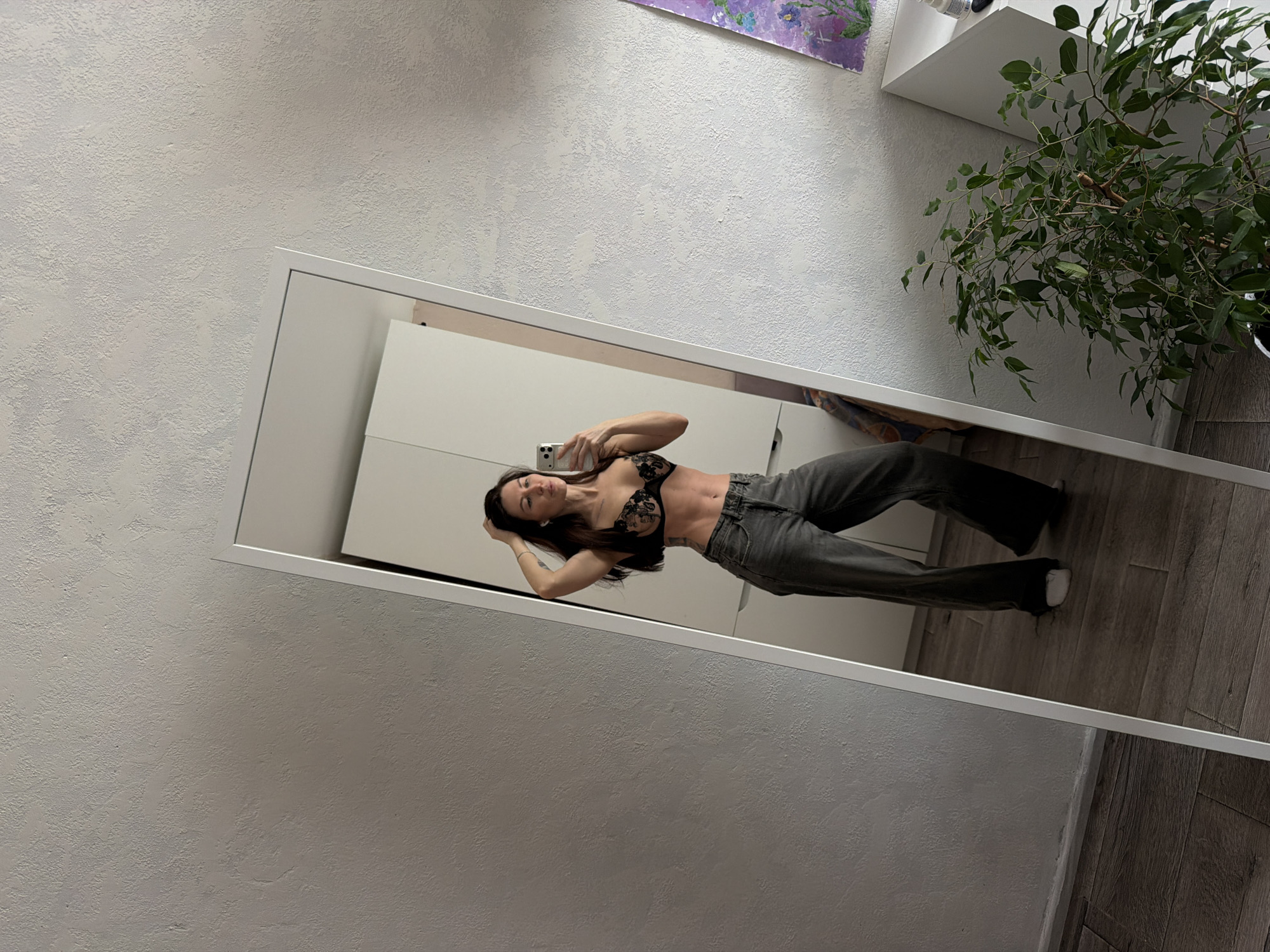 The image captures a woman taking a mirror selfie in an indoor setting. She is reflected in a white-framed full-length mirror, visible from her chest down to her feet. The woman is wearing a black lace bralette and dark gray jeans, with white socks. Her left hand holds a white smartphone up to capture the image, while her right hand is positioned behind her head, resting on her dark hair. Her toned midsection is prominently displayed.
The immediate surroundings of the mirror reveal a light grey wall with a distinct rough, textured finish. To the left and above the mirror's frame, this textured wall is visible. Above the mirror, on the textured wall, there is a colorful abstract painting featuring shades of purple and green, with a white wall-mounted shelf beneath it. To the far right of the image, near the top, part of a green leafy plant is visible. The floor, visible outside the mirror frame and reflecting the woman's lower legs, consists of wooden planks. The lighting suggests an indoor environment. The woman's expression appears neutral as she focuses on her phone. No visible text is present in the image.