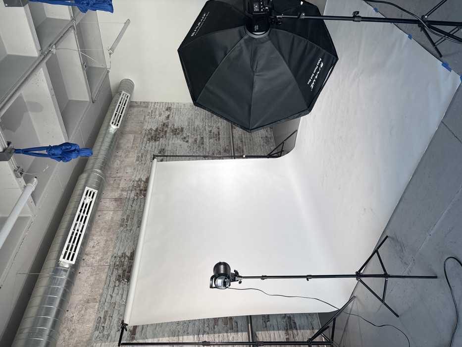 This image depicts a photography studio located in Unknown, Unknown. A large white seamless paper backdrop is deployed from a stand across the back of the room, extending onto a light gray concrete floor where it is secured with blue adhesive tape. Two professional studio lights are set up on black tripod stands. On the left, a studio strobe with a silver reflector is aimed at the backdrop. On the right, a large black octagonal softbox, labeled 'Godox P90L Parabolic Softbox', is mounted on a strobe, also directed towards the backdrop. The walls behind the backdrop consist of exposed red brick on the left and a plain white painted surface on the right. The ceiling features exposed silver HVAC ductwork with multiple grilles, and two bundles of blue fabric slings are suspended from ceiling mounts. Electrical cords run along the floor. No individuals are present in the scene.