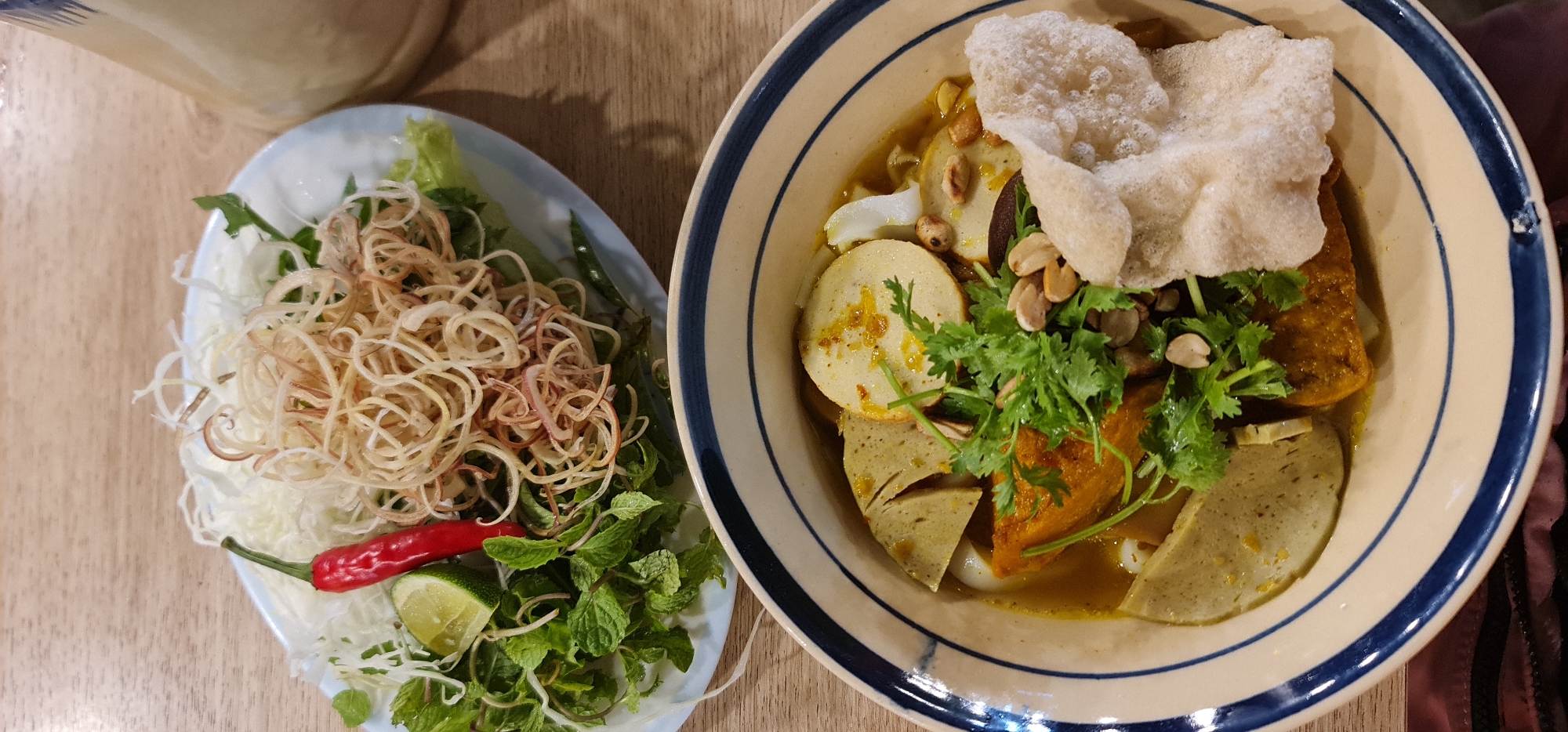 A high-angle, medium shot depicts a Vietnamese meal, specifically mì quảng, on a wooden table. A light blue, oval plate contains a garnish of shredded banana blossom, green herbs including mint, a red chili pepper, and a lime wedge. Beside it is a bowl with a tan rim and blue lines. Inside the bowl is the mì quảng. Elements visible include turmeric-infused broth, rice noodles, pork sausage, peanuts, fresh coriander, and rice crackers. The meal is presented indoors, possibly at a restaurant in Đà Nẵng, Vietnam.