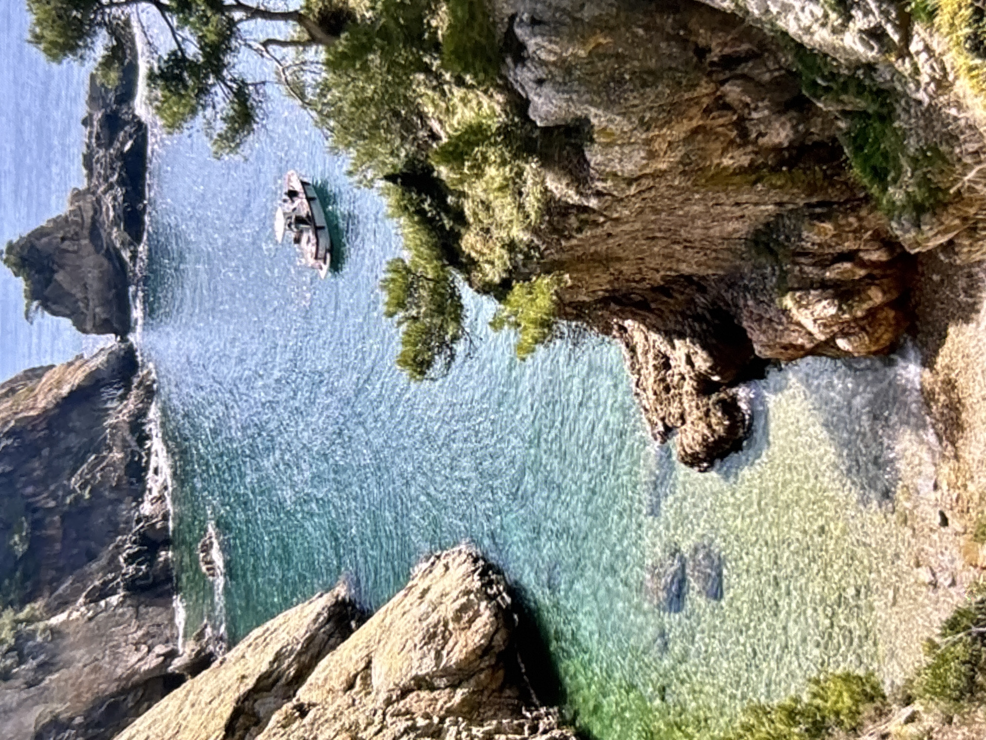This image depicts a picturesque cove with crystal-clear turquoise water, flanked by rugged, sun-drenched cliffs. A small white boat with several people onboard navigates the calm waters, leaving a gentle wake. Lush green vegetation clings to the rocky outcrops, adding a vibrant contrast to the earthy tones of the cliffs and the blue of the sea. The seabed is visible through the shallow water near the shore, revealing submerged rocks. The scene suggests a tranquil day in a Mediterranean coastal location, likely a natural park or a protected area, potentially near the coast of Orihuela, Spain, known for its dramatic coastal landscapes.
