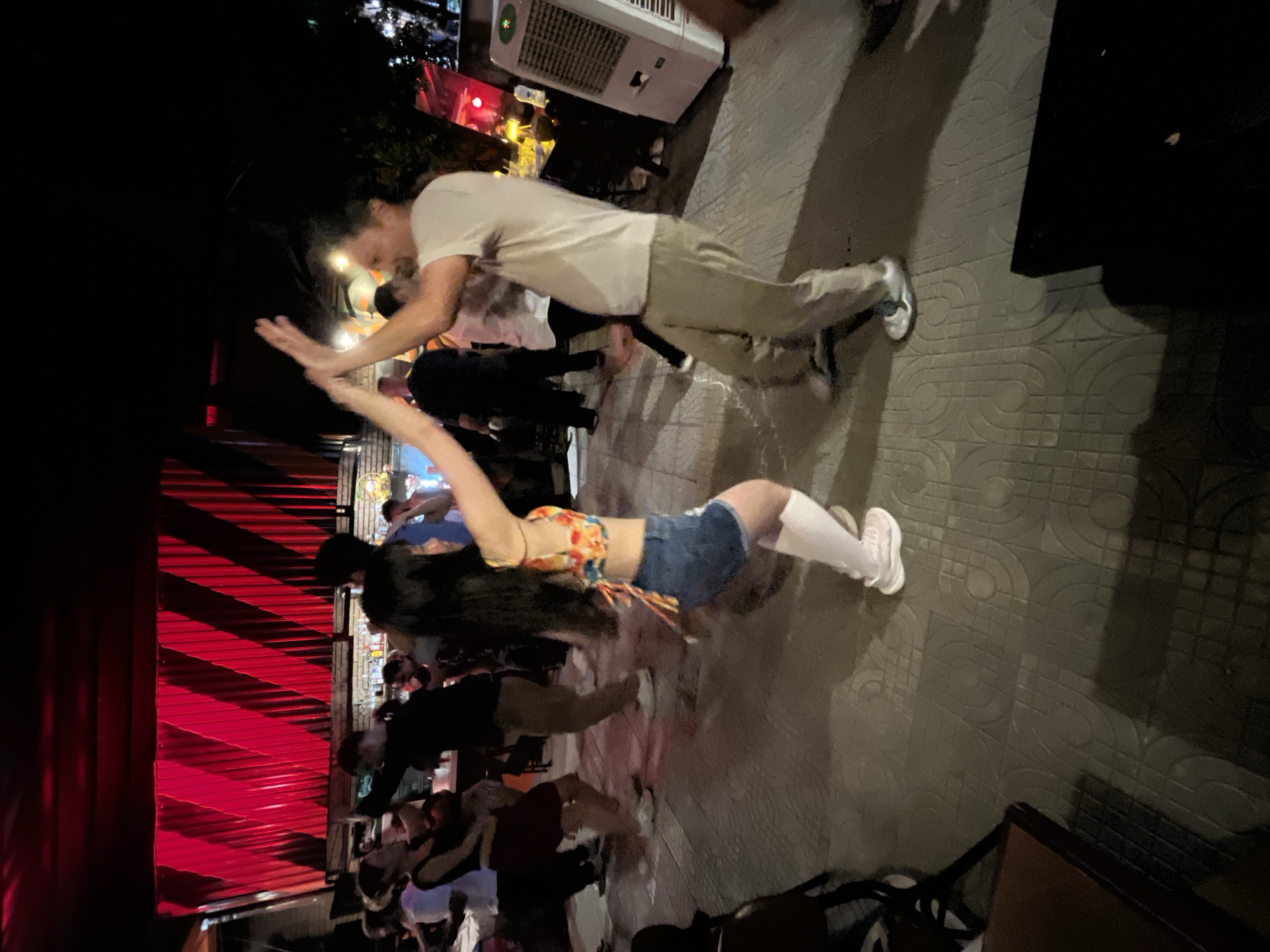 This image depicts a nighttime scene at an outdoor or semi-outdoor social venue in Da Nang, Vietnam, where approximately eight to ten individuals are visible, primarily engaged in dancing. In the central foreground, a woman with long dark hair, clad in a colorful floral crop top, blue denim shorts, and white sneakers with long white socks, is actively dancing. Her hands are raised and connected with those of a man to her right, who is wearing a light-colored t-shirt, light-colored trousers, and white sneakers; both individuals are in motion, suggesting a partner dance.
Behind the central dancers, multiple other people are visible, some in various stages of dancing or standing near a bar area where bottles are visible. The background is dominated by a prominent wall featuring red corrugated panels arranged with diagonal stripes, illuminated by red lighting. To the far right, a white, upright air cooler or large fan is present. The ground is paved with light-colored tiles exhibiting a repetitive curvilinear pattern. Dark-colored furniture, including a table and chairs, is partially visible in the lower left foreground. The ambient lighting is dim, characteristic of a nightlife setting, with focused red illumination on the background wall.