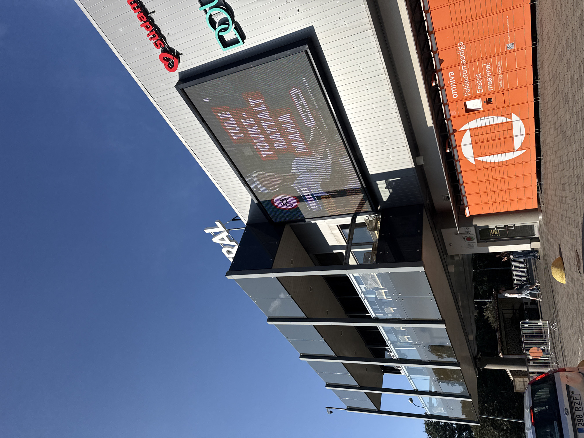 This is an exterior, low-angle shot of a modern building with a bright blue sky. The building features a large digital screen displaying text and an image of a cyclist. The text on the screen appears to be in Estonian and reads, "TULE TOUKE-RATTALT MAHA" which translates to "Come off the spring bike." Below this, there is smaller text that includes a website address. To the right of the screen, a teal logo with the letters "nou" is visible.
Above the digital screen, a red neon sign with a heart symbol and the word "sudame" is mounted on the building's facade, suggesting a connection to a retail or service establishment. The building's exterior is predominantly silver-colored, with dark grey structural elements and glass balconies on the upper levels.
In the foreground, an orange parcel locker station, identified by the "omniva" logo and text in Estonian, is prominently displayed. The text on the locker station reads, "Paki automaadiga Eestist maailma!" which translates to "With the parcel machine from Estonia to the world!"
A silver Ford vehicle with a license plate reading "298 RZF" is parked on the left side of the frame. Several people are visible walking in the middle ground near the building's entrance. The environment appears to be an urban setting, likely in Tallinn, Estonia, given the presence of Estonian text and the "omniva" branding, which is a Lithuanian postal and logistics company that also operates in Estonia.