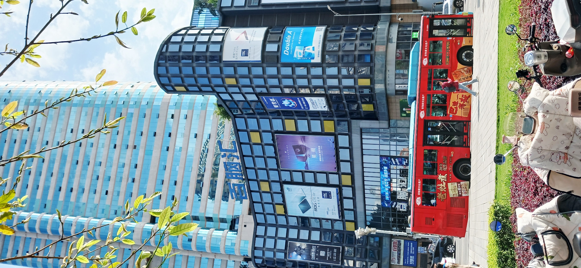 This is an outdoor shot of a city street, taken in the Jiulongpo District of Chongqing, China. It features a tall building with blue and white vertical panels and a unique curved architectural design that incorporates a grid-like facade. Advertising panels are visible on the building, with brands such as "Double A" and "ROG."
A red bus is parked on the right side of the image, and a pedestrian is seen stepping onto the sidewalk from the bus. Further right, there are parked mopeds with protective covers, positioned near a flowerbed.
In the foreground, a tree branch with new leaves frames the scene, adding a touch of nature. The sky is visible in the background with light clouds, indicating a bright day.