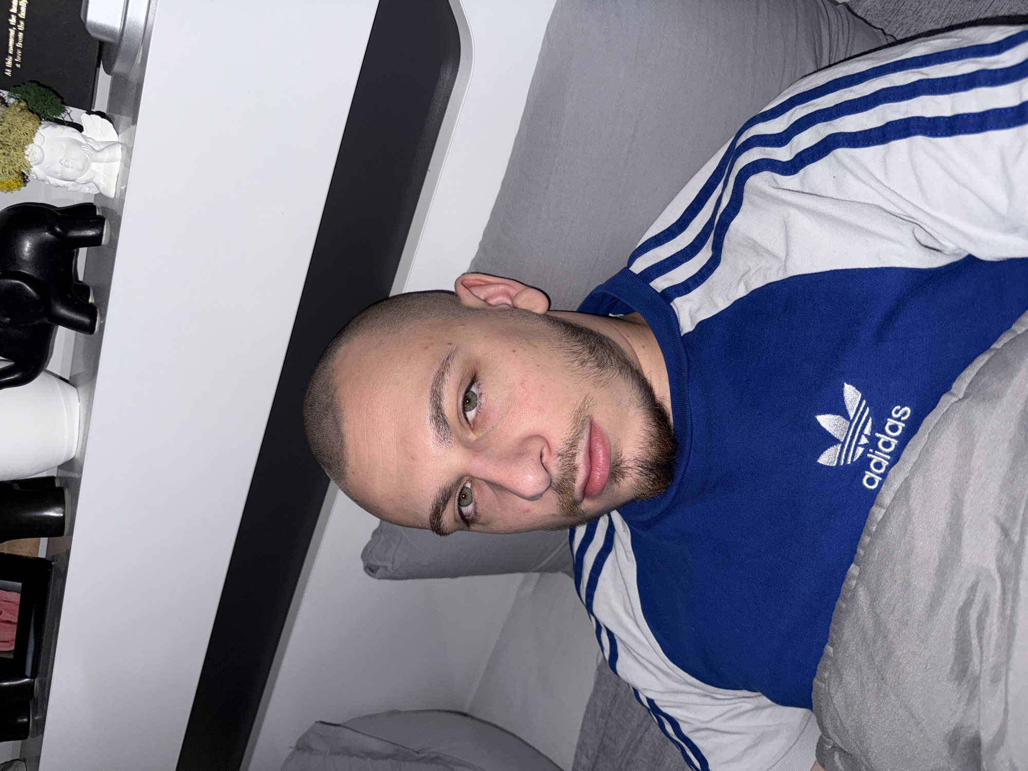 A man with a shaved head and beard is lying in bed, wearing a blue and white Adidas shirt. He is facing the camera. The background includes a white and black shelf unit with various decorative items, including a black elephant figurine, a white statue with moss, and a silver container. The environment suggests a residential bedroom setting. The location in Stara Zagora, Bulgaria, cannot be determined from the provided image.