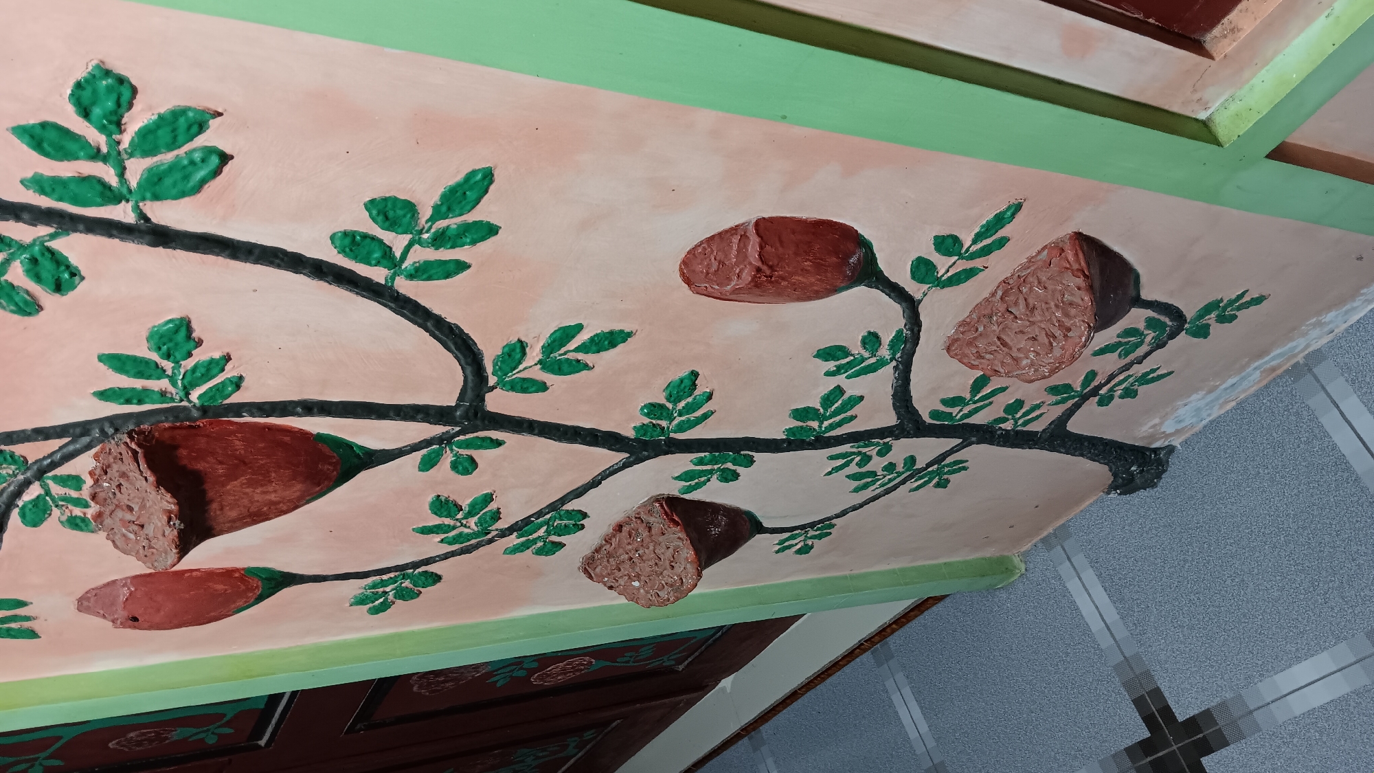 The image depicts a textured mural on a pale orange wall, likely located in Sonobekel, Indonesia. The mural features a stylized black tree branch with green leaves, and several red, bud-like structures that appear to be either flowers or fruits. Some of these red elements are textured, resembling opened seed pods or fruits with rough interiors. The mural is bordered by a light green frame on the right and a dark wooden door on the left. The floor in the foreground is a gray speckled tile with a grid pattern. The scene appears to be an exterior wall or hallway. There are no people or animals visible in the image.