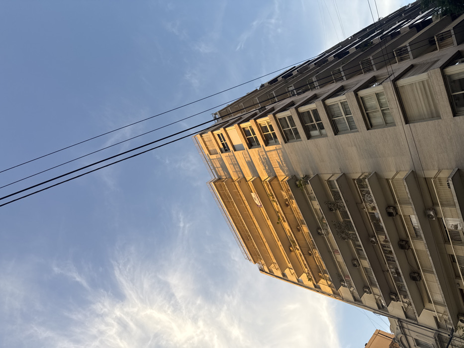 An upward-angle photograph captures a multi-story residential apartment building during late afternoon, evident by the warm, golden sunlight illuminating its left facade. The sky is clear blue with wispy white clouds. Four black utility lines traverse the upper right quadrant of the frame diagonally.
The building, constructed of light-colored, possibly beige, material, presents at least twelve visible floors. The sunlit left facade features numerous balconies with white railings. These balconies house approximately 15-20 external air conditioning units, various potted plants, and small trees. One lower balcony holds a white outdoor table and two chairs. A small, unreadable white sign is attached to an upper balcony railing. The building's right facade is partially shadowed, displaying multiple windows, some equipped with external roll-down shutters. A portion of an older, darker-toned building is visible in the lower-left background. No people are present. The architectural style, prevalent balconies, visible air conditioning units, and overhead utility lines are characteristic of a typical residential street within the city of Buenos Aires, Argentina.