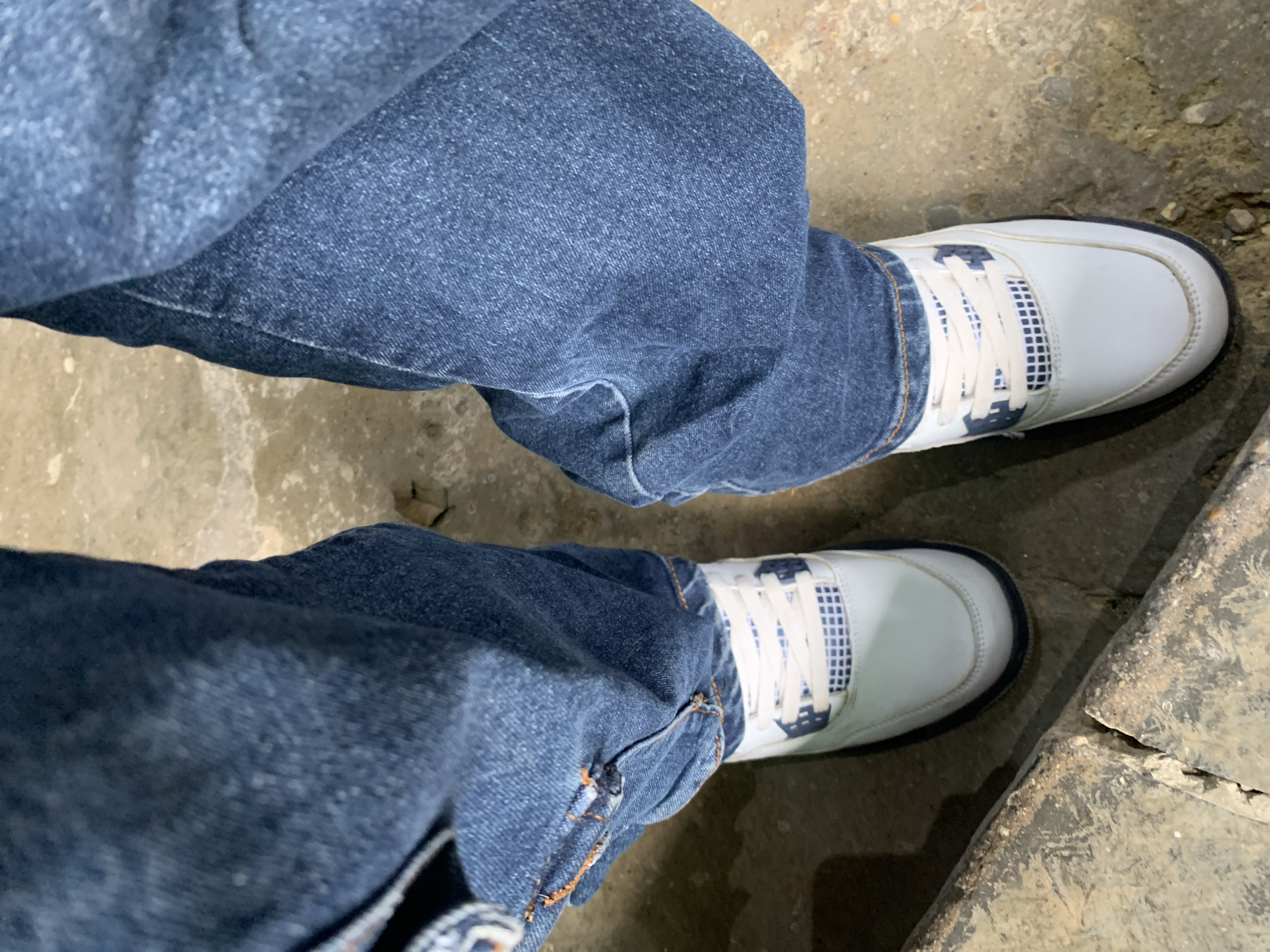 The image captures a top-down view of a person's lower body, standing still. The individual is wearing dark blue, relaxed-fit jeans, which are slightly bunched around the ankles, partially covering their shoes. On their feet are white sneakers with distinct blue accents, including a blue toe cap and a blue and white grid pattern visible on the side panels and around the laces. The laces are white.
The person is standing on a rough, uneven ground surface, which appears to be concrete or asphalt. The ground exhibits varying shades of gray and brown, with textures suggesting wear and tear, and some small debris or gravel visible. Towards the right side of the frame, the ground becomes darker and more fractured, with a noticeable crack running across. The lighting is diffused, making the exact time of day or weather conditions unclear. The scene provides a glimpse of an outdoor or utilitarian indoor setting in La Tagua, Venezuela. No visible text is present in the image.