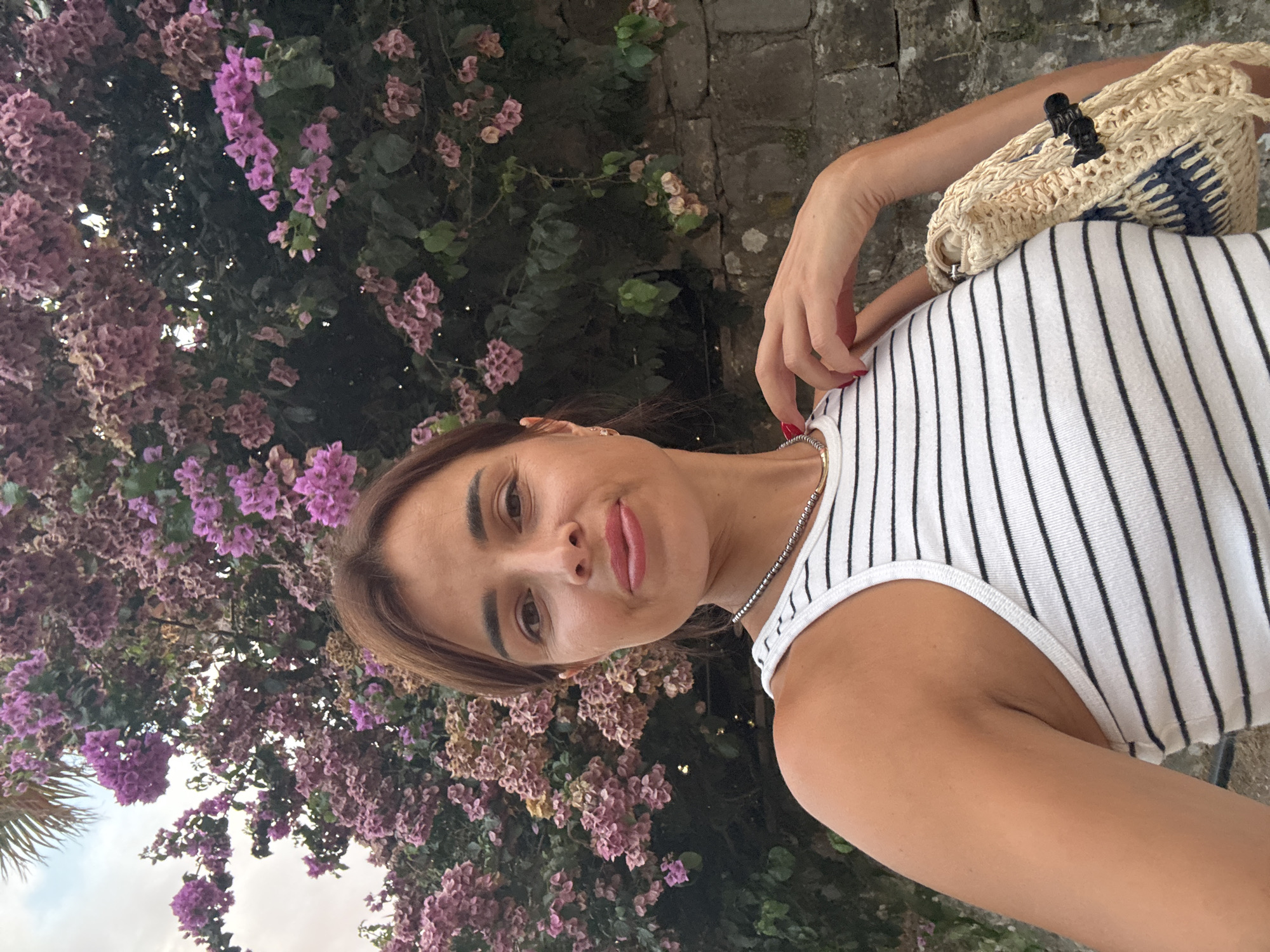 A selfie taken by a woman standing outdoors with a wall of bougainvillea flowers behind her. She is facing the camera and smiling slightly, with her right hand touching her neck. She is wearing a white and black striped tank top and a necklace. A straw bag is visible on her right side. The background consists of lush pink and purple bougainvillea flowers, green leaves, a stone wall, and a palm frond in the upper left corner.