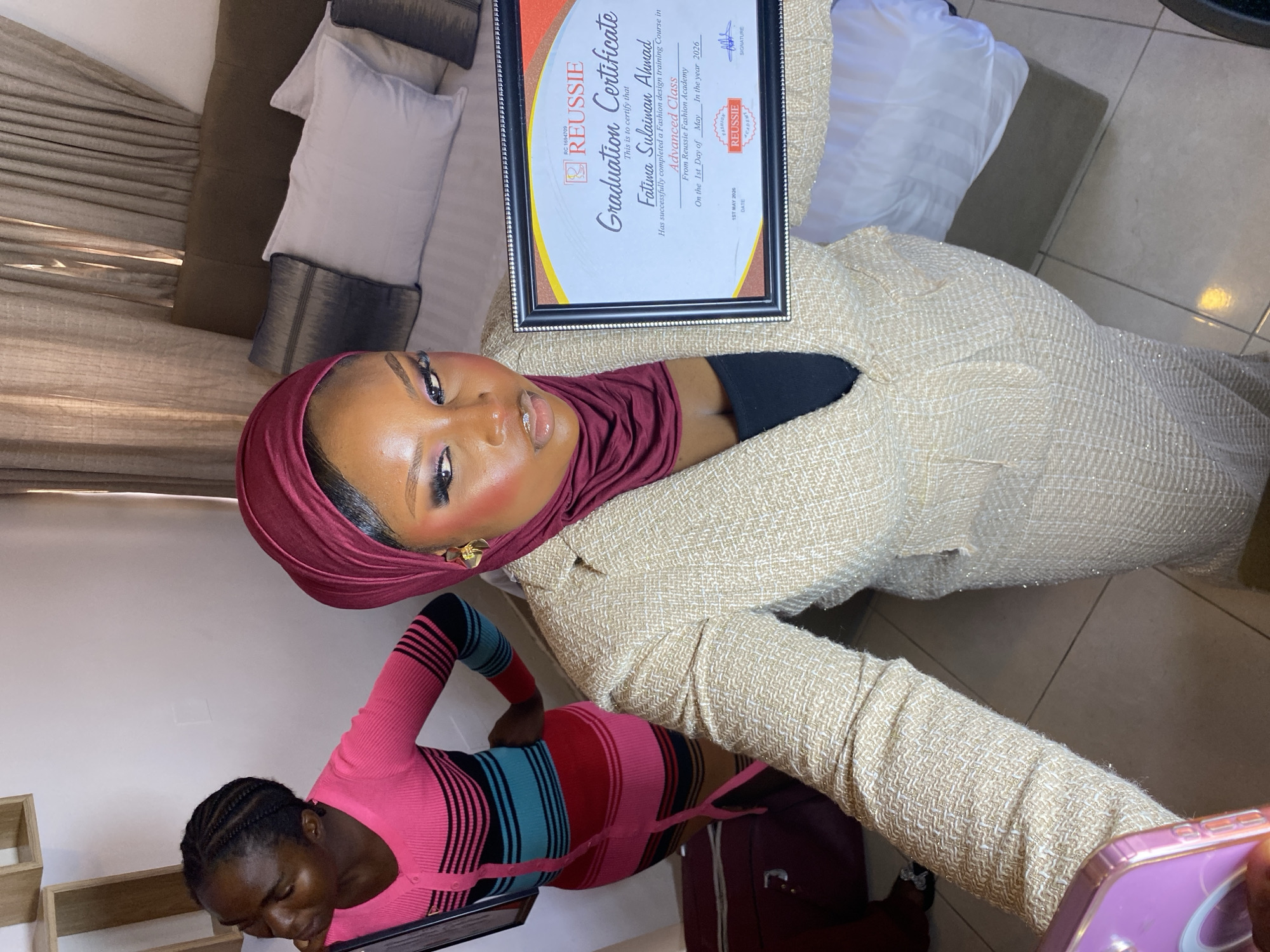 An indoor, vertically oriented image, showing two women in what appears to be a home or studio setting in Gura Top, Nigeria. The main subject, a woman with a maroon hijab and a light-colored, textured dress or top, is positioned in the upper right. She is proudly holding up a framed "Graduation Certificate." Her face, adorned with visible makeup, is turned towards the viewer.
The certificate, from "REUSSIE Fashion Academy," is awarded to "Fatima Sulaiman Ahmad" for successfully completing an "Advanced Class" in Fashion design training. The date on the certificate reads "1ST MAY 2026." Text visible on the certificate also includes "RC 1694709," "SIGNATURE," and "FASHION ACADEMY."
In the lower left, another woman with braided hair, wearing a striped pink, blue, and black top, is visible, holding a smartphone, presumably taking the picture or video of the main subject.
The background features light-colored pillows and bedding, suggesting a couch or bed, along with a tiled floor. Wooden shelves or boxes are partially visible on the left. A dark-colored piece of luggage or bag is seen on the right. The overall impression is one of celebration and achievement, captured in a personal setting.