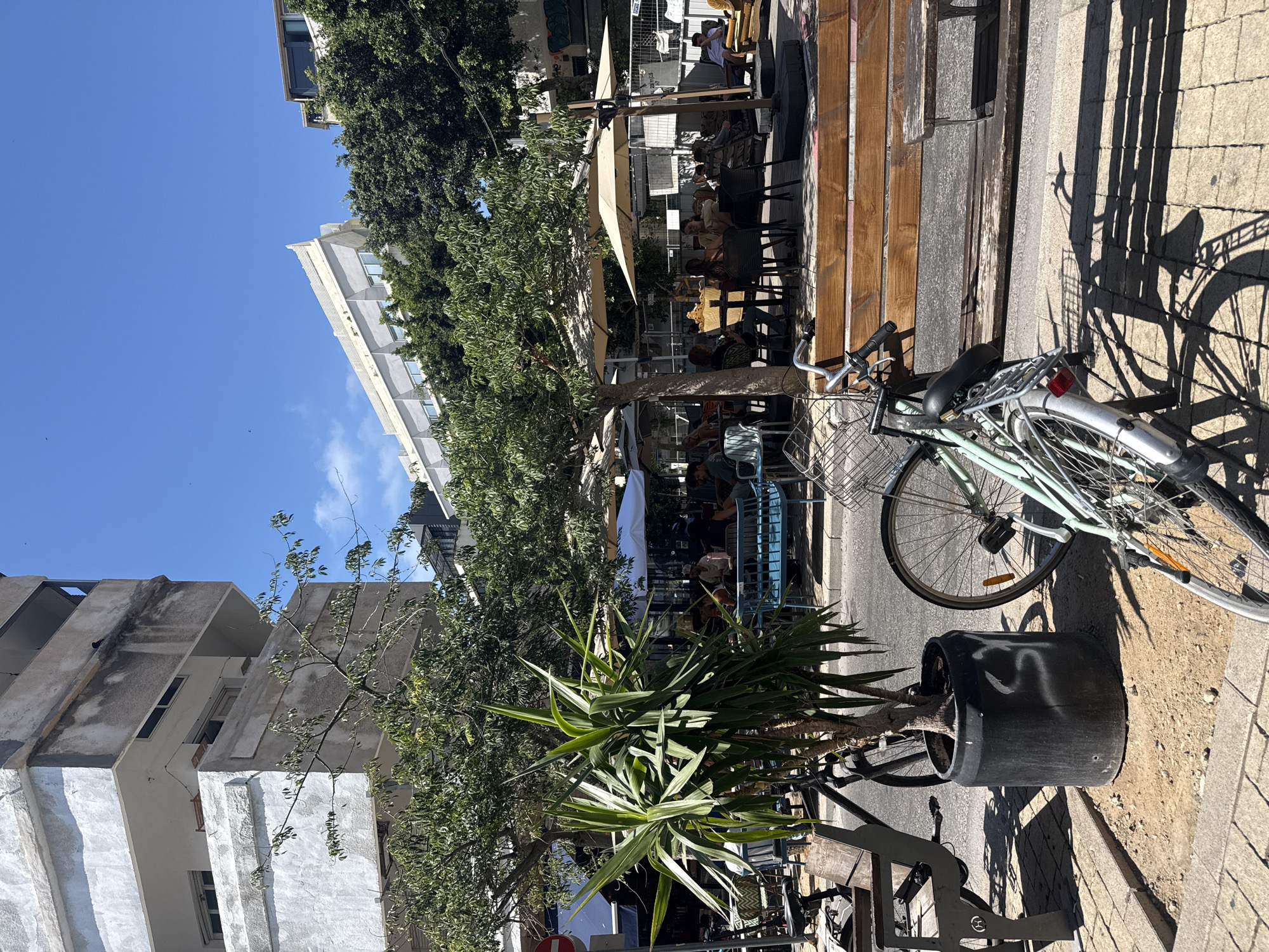 The image shows an outdoor cafe scene in Tel Aviv, Israel. People are seated at tables under umbrellas. A light blue bicycle is parked on the sidewalk near a large potted plant. Buildings are visible in the background, and the sky is blue.