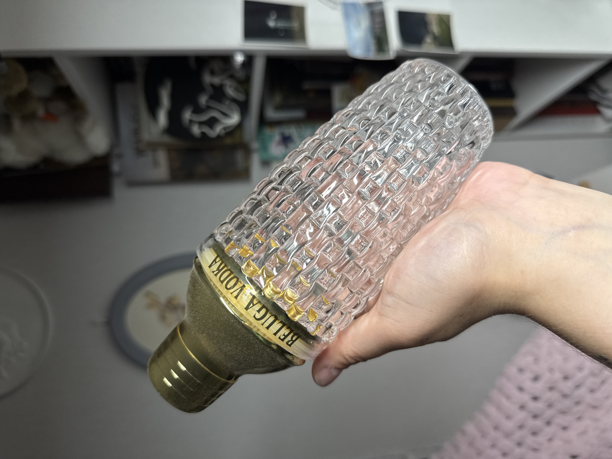 A human hand with visible light skin and subtle veining on the wrist is holding a clear glass cocktail shaker. The shaker features a distinct basket-weave texture across its body and is topped with a frosted gold-colored lid. A yellow label encircles the upper portion of the shaker, prominently displaying "BELUGA VODKA" in gold, raised lettering. In the blurred background, a soft pink, likely knitted or crocheted, textile is visible on the lower left. Centered on the wall is a light-colored, framed circular decorative item. To the right, a white shelving unit is visible, holding various items: amorphous, light-colored objects on the top shelf, a dark item with white, stylized mushroom shapes, and several small framed pictures or postcards showing landscapes and abstract designs on lower shelves. The scene is set indoors, within what appears to be a personal or residential environment.
