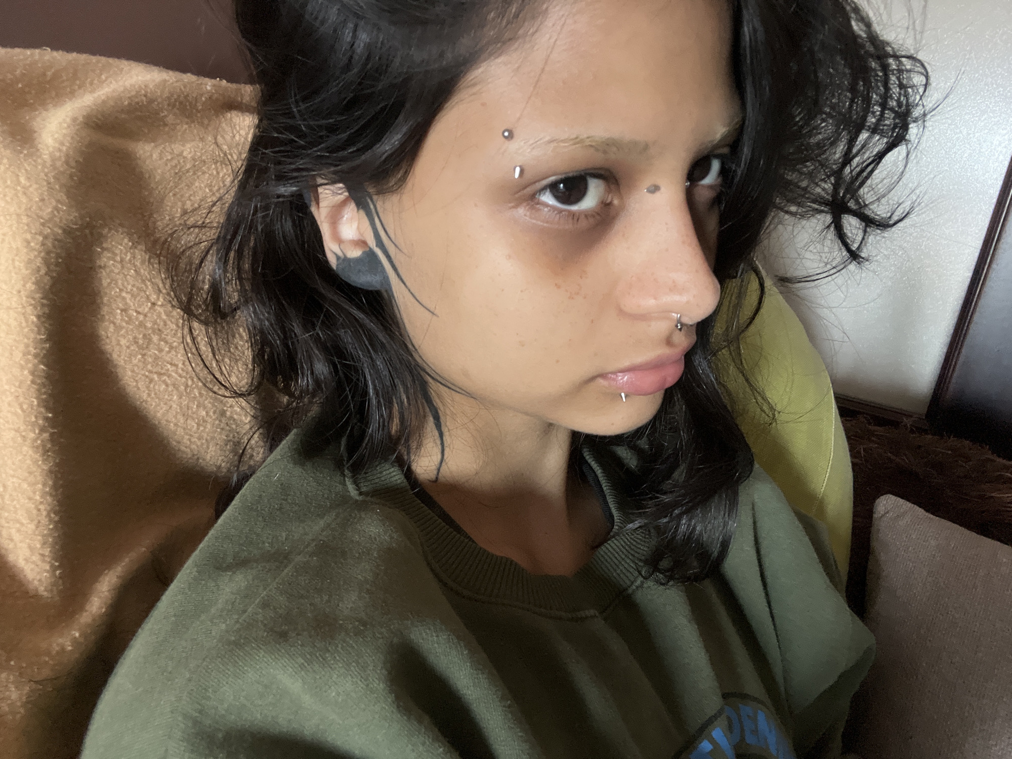 A close-up shot depicts a person with dark hair and fair skin. They have multiple piercings: two on the left eyebrow, one on the right cheek, a nose ring, and a lip ring. The left ear features a dark tattoo. They are wearing a green sweatshirt and are seated on a brown and beige surface. The location, Arroyo De La Miel-benalmadena Costa, Spain, is not evident in the image.