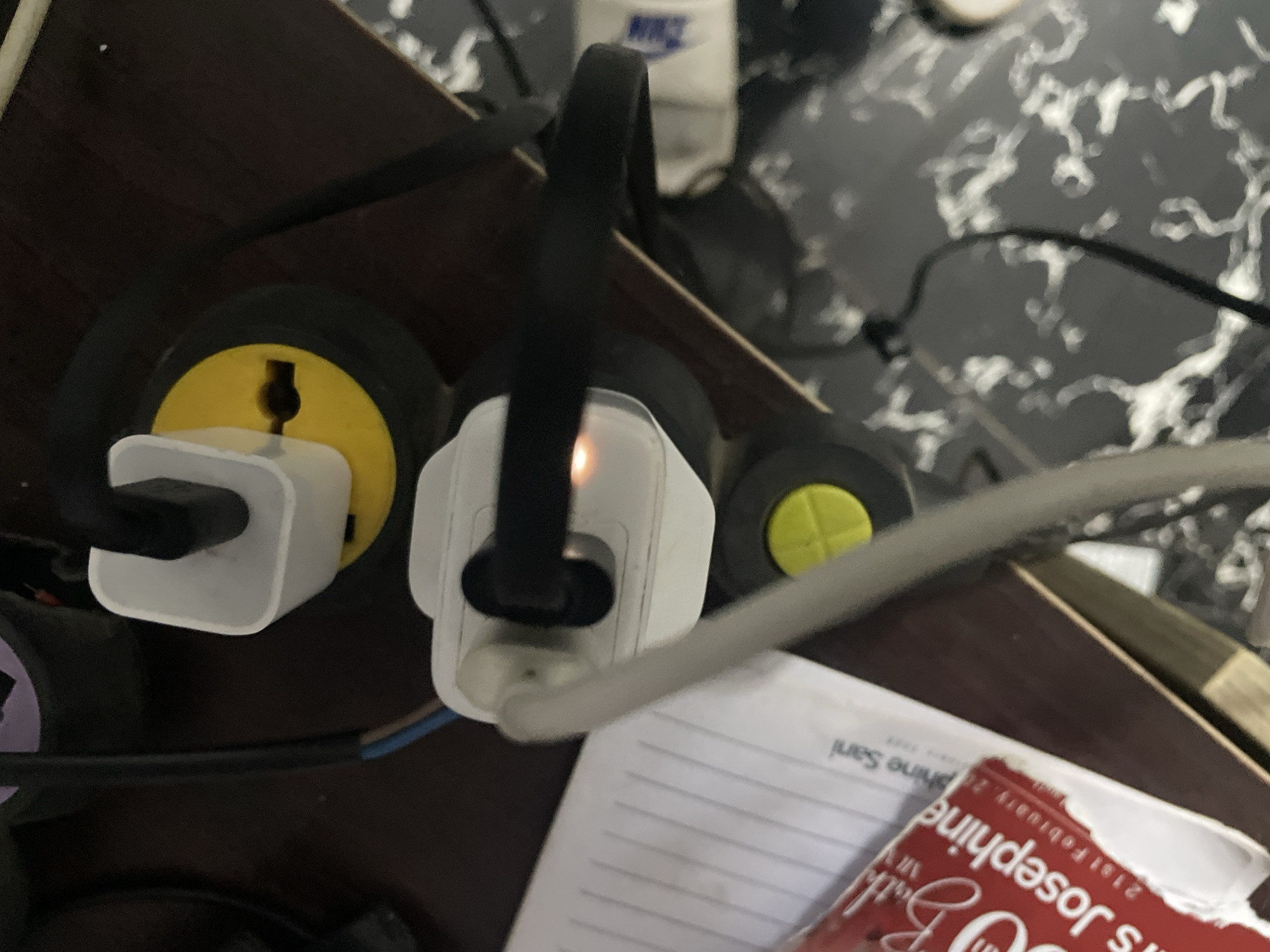 The image presents a close-up, downward view of a power strip and associated electrical accessories, situated on a dark wooden surface with a patterned dark background. At the core, a black power strip features several outlets. One yellow-accented universal adapter is plugged into an outlet, housing a white USB wall charger. Adjacent to it, another white USB wall charger is directly plugged into the power strip, emitting an orange glow, signifying its active state, likely charging a device. Multiple black and grey cables extend from these chargers, and a blue and brown electrical wire is also visible.
In the upper background, a dark surface with a light, swirly pattern, resembling marble, is seen. Partially visible behind the power strip is a white object with the blue "NIKE" logo. To the lower right, a white sheet of lined paper, possibly from a notebook, shows inverted text including "Sani" and "21st February." Overlapping this paper is a red item, possibly a book cover or box, also displaying inverted white text: "My Beloved," "Mrs Josephine," and "21st February." The scene depicts an indoor setting, such as a home or office in Abuja, Nigeria, where electronic devices are being powered or charged.