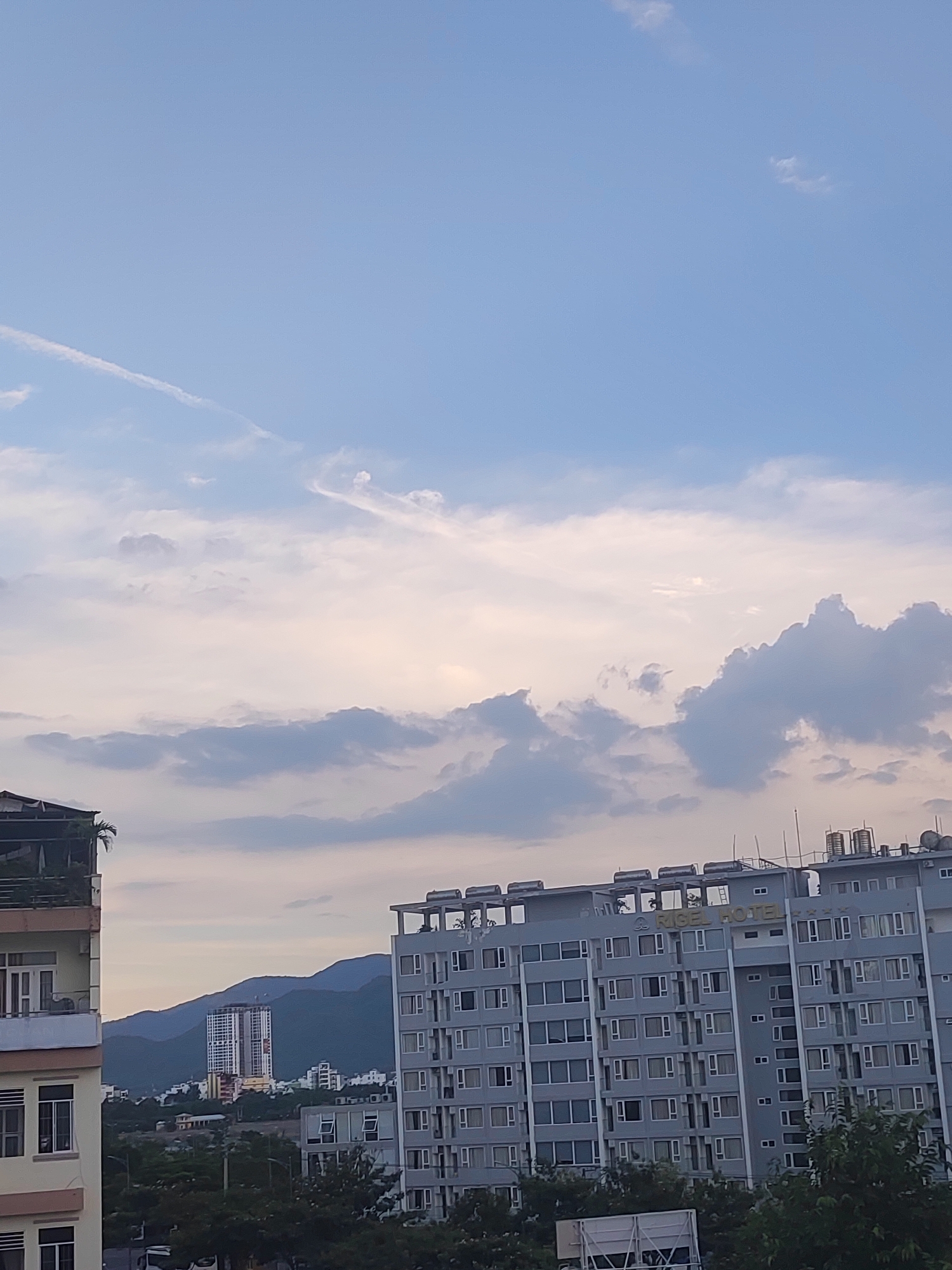 The image shows a view of a city with buildings and mountains in the background, under a partly cloudy sky. In the foreground, there is a multi-story apartment building on the left and a large hotel building on the right. The hotel building has the name "RIGEL HOTEL" visible in gold lettering on its facade. Several solar water heaters are visible on the rooftops of the hotel and another building. In the middle ground, there is a tall, unfinished building under construction, situated between a range of dark green, undulating mountains. A few smaller buildings and trees are scattered between the foreground structures and the mountains. The sky is a pale blue with wispy white clouds and some larger, darker clouds in the middle and upper portions of the image. The scene appears to be captured during the late afternoon or early evening, given the lighting conditions. The location is identified as Nha Trang Ward, Vietnam. There are no people visible in the image.