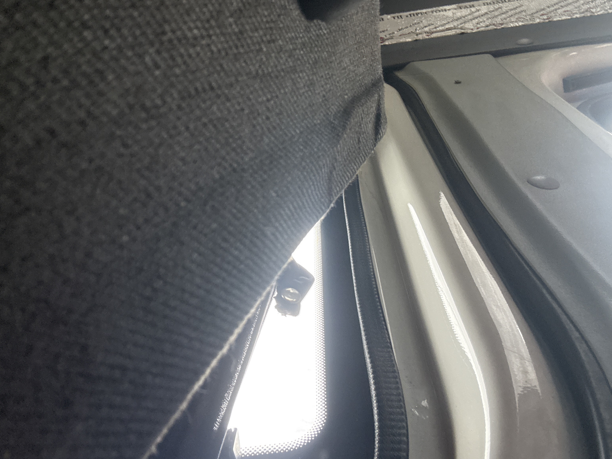 The image shows a close-up, low-angle view of the interior of a vehicle, likely a van or SUV, with a focus on the ceiling and upper interior panels. The dominant elements are the dark grey fabric headliner, which occupies the upper half of the frame and shows a textured weave pattern, and the white or light grey interior paneling below. A bright light source, likely sunlight through a windshield or sunroof, illuminates a horizontal band across the middle of the image, creating a glare. Attached to the windshield, within the illuminated area, is a small black electronic device, possibly a dashcam or sensor, with a visible lens. A black rubber seal or trim piece runs horizontally along the edge where the headliner meets the upper interior panels. The lower portion of the image reveals curved white panels with some subtle reflections and visible mounting points. To the far right, a section of silver, textured insulation material is visible, partially obscured by the interior paneling. There are no people or overt actions depicted. The scene suggests the interior of a parked vehicle, the specific location within Mahilyow, Belarus, cannot be determined from the image alone.