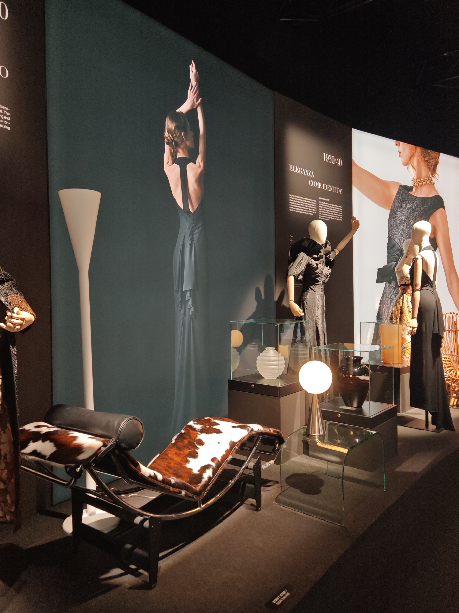 This media file captures an indoor exhibition space, likely within a museum or gallery, dedicated to fashion and design from the 1930s and 1940s. The setting is dimly lit, with strategic lighting illuminating the exhibits against dark walls and a dark floor.
No real people are present; the display features two wooden-jointed mannequins. One on the left wears a dark, possibly black, dress with dramatic puffed and ruched sleeves, while the other on the right sports a flowing black gown with an open back. Large photographic wall panels complement the garments, one showing a woman from behind with raised arms in an elegant backless dress, and another presenting a woman's profile wearing a patterned top and a prominent gold chain necklace.
The exhibition includes notable furniture pieces and decorative objects. A distinctive Le Corbusier LC4 chaise lounge, featuring a black frame and striking brown and white cowhide-patterned upholstery, with a black cylindrical headrest, occupies the foreground. Beside it, a clear, bent-glass side table holds a modern spherical white table lamp. Other lighting includes a tall, slender white floor lamp. Various decorative items, such as a frosted ribbed vase and a dark ceramic vase, are displayed within clear glass cases set on black pedestals. A small wicker chair is partially visible in the background.
The visible text on the wall panels reads "1930/40" and "ELEGANZA COME IDENTITA'" (Elegance as Identity), indicating the exhibition's theme and period. A small warning sticker on the floor states "DON'T STEP / NON SALIRE" (Do not step / Do not climb). The atmosphere is carefully curated, aiming to present historical fashion and design in an elegant context. The location context is unknown.