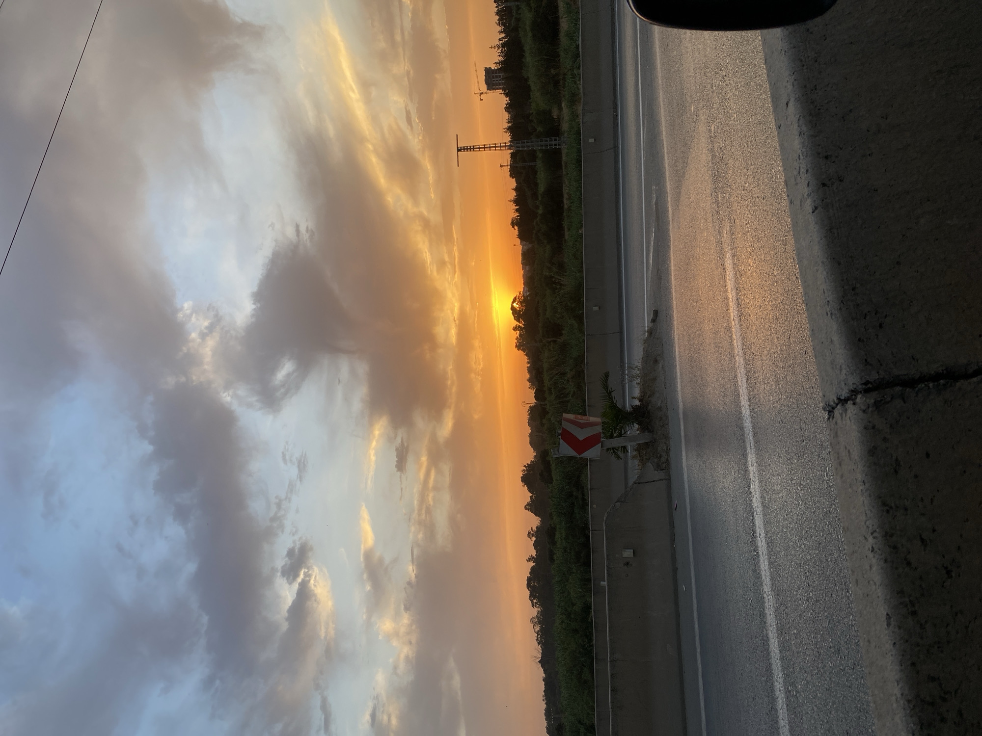 The image shows a scenic view of a highway at sunset in El Karia, Algeria. The sky is filled with dramatic clouds tinged with orange and yellow hues from the setting sun, which is partially obscured by trees on the horizon. A concrete barrier runs along the side of the highway, separating it from a strip of green vegetation. A traffic sign with red and white chevrons indicating a curve is visible on the left side of the road. In the background, there are more trees and a few structures, including what appears to be a tall tower or mast and a building with a crane nearby, suggesting industrial or construction activity. The asphalt road is wet and reflects the warm colors of the sky. The overall scene captures a tranquil moment of a sunset over a highway environment.
