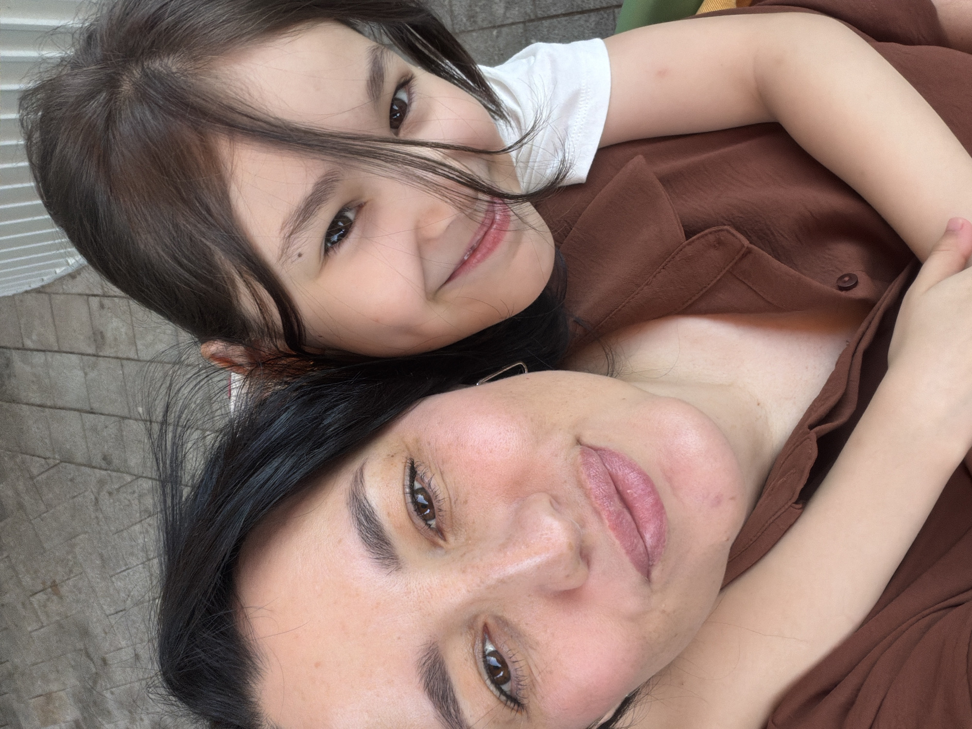 In this close-up, high-angle selfie, a woman and a young girl are embracing outdoors. The woman, on the left, has dark hair and fair skin with freckles. She is wearing a brown shirt. The girl, on the right, has brown hair and fair skin. She is wearing a white shirt. They appear to be in a public space, possibly a park or a street, given the stone-like texture in the background. The lighting suggests it is daytime. The overall mood is affectionate, with both individuals smiling and looking towards the camera. There is no discernible information to place this image specifically in Moscow, Russia.