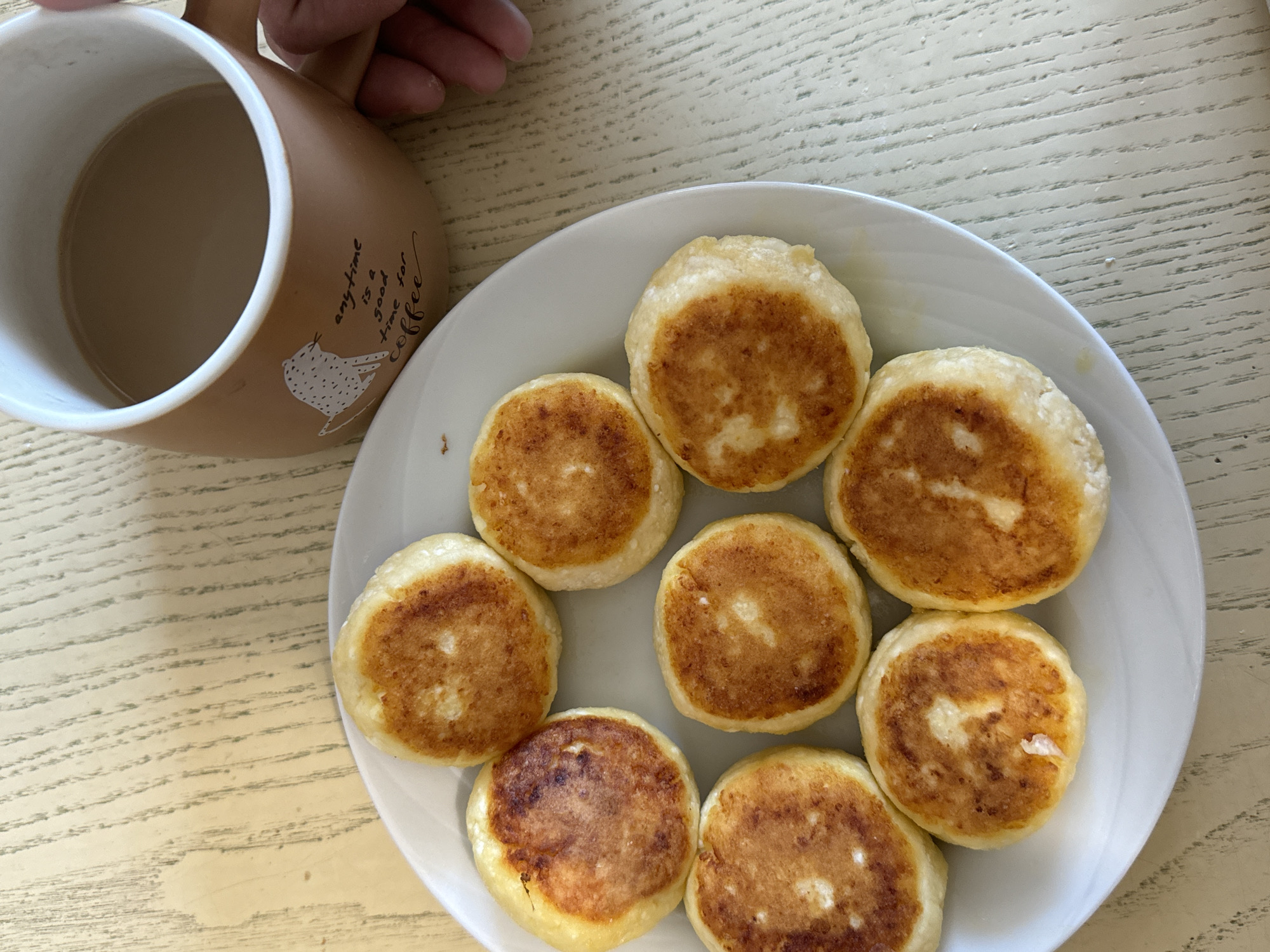 A high-angle shot depicts a table scene with breakfast. A white plate containing eight small, round, golden-brown pancakes arranged in a circular pattern occupies the right side of the frame. Adjacent to the plate, on the left, is a brown mug filled with a light-colored beverage, likely coffee or tea with milk. A person's hand is visible near the mug. The table has a light-colored, textured surface. The image was taken in a Chaikovsky (Russia) residential dwelling.