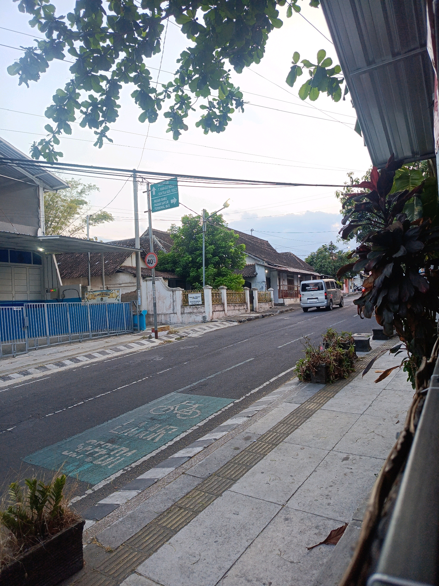 The image depicts a street scene in Magetan, Indonesia, likely in the early morning or late afternoon due to the soft lighting and diffused shadows. A road with painted lane markings runs through the center, bordered by sidewalks. On the left side of the road, there is a building with a blue metal fence and a corrugated metal awning. A street signpost with directional arrows and text in Indonesian is visible, along with a circular speed limit sign indicating "40". A silver van is driving away from the camera on the right side of the road. The sidewalks are made of concrete tiles, with tactile paving for the visually impaired. Planters with sparse vegetation are placed along the sidewalk. Trees with green leaves overhang the scene from the top left, and a darker-leaved plant is visible on the right side of the frame, partially obscuring a metal structure. The sky is overcast with scattered clouds. There are no people clearly visible in the image.