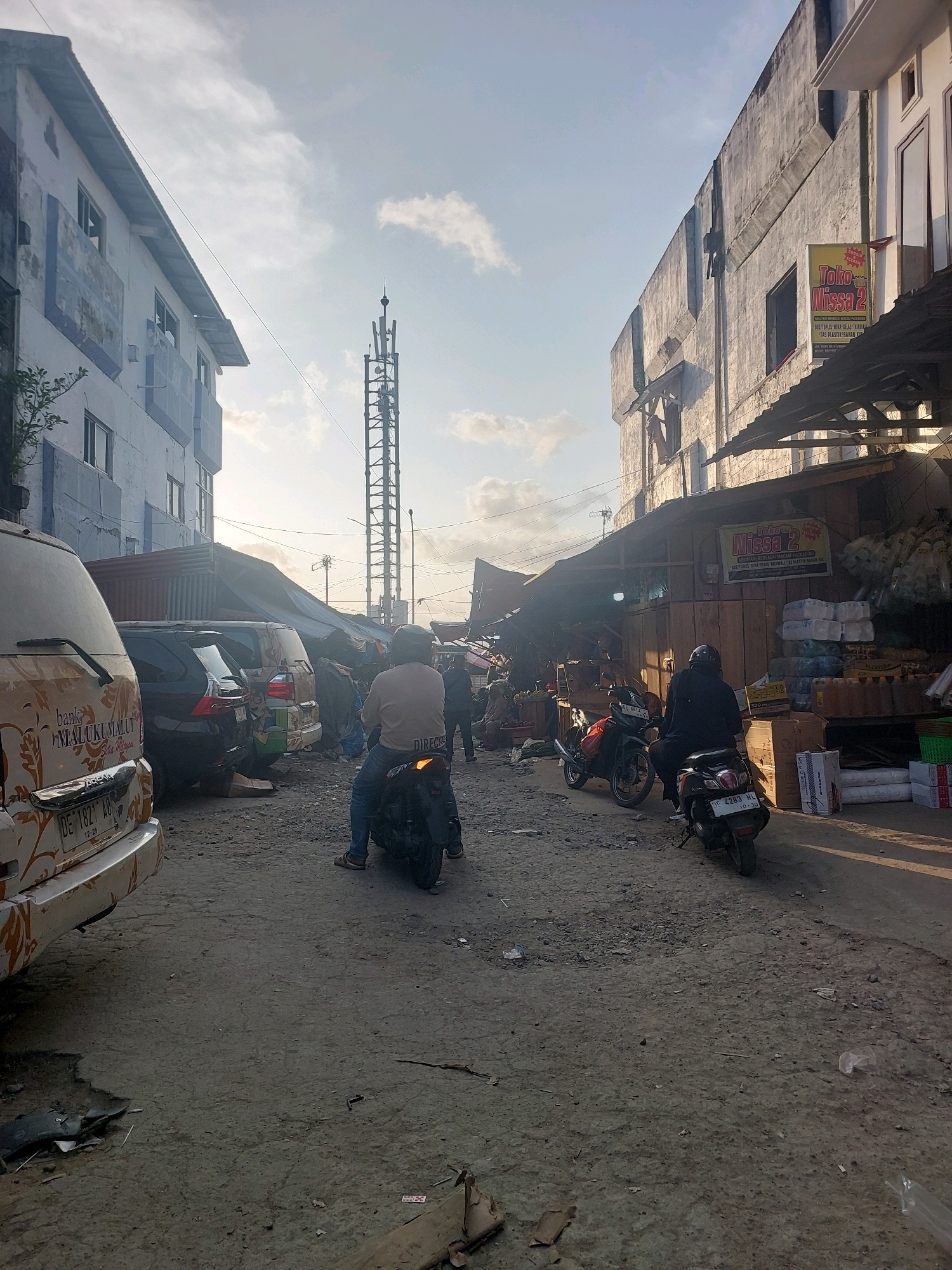 The image depicts a street scene in a commercial area, likely a market, in Ambon, Indonesia. The foreground is dominated by a rough, unpaved road with scattered debris. Several vehicles are present, including a white van with an ornate design on its side, a black SUV, and two motorcycles. One motorcycle, ridden by a person wearing a light-colored shirt and jeans, is positioned in the center of the frame. Another motorcycle, with a rider in a black outfit, is further back on the right.
Buildings line both sides of the street. On the left, a multi-story building with a light-colored facade and blue accents is visible. On the right, a long, weathered building with a corrugated metal roof forms a market stall area. Several stacks of goods, including plastic containers and boxes, are piled along the storefront. A prominent sign with red text reading "Toko Nissa 2" hangs above one of the stalls. In the background, a tall telecommunications tower stands against a partly cloudy sky. The overall impression is a busy, functional urban environment.