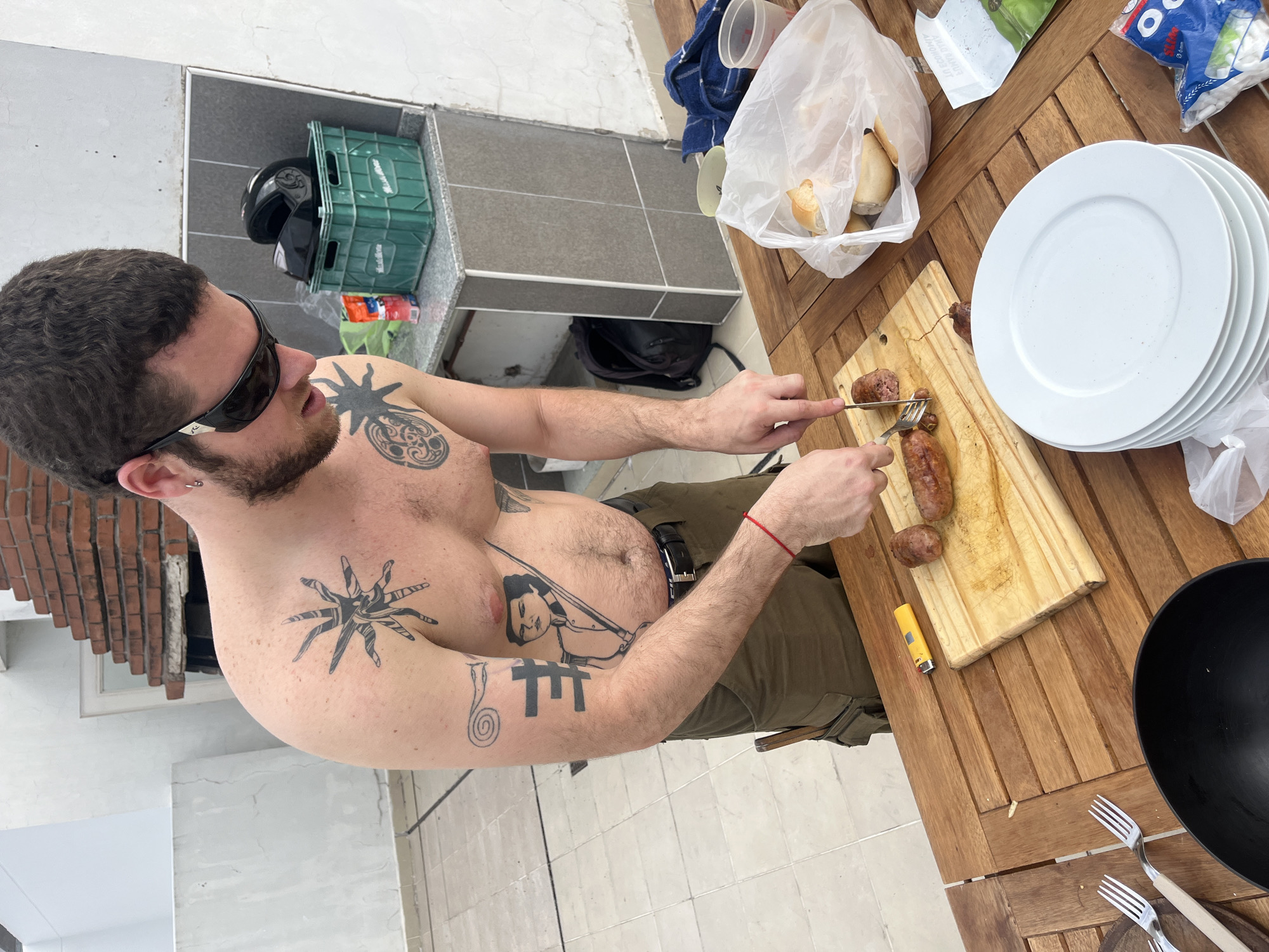 A shirtless man with tattoos is cutting sausages on a wooden cutting board at a wooden table. He is wearing sunglasses and green pants with a black belt. On the table are plates, bread, forks, a bowl, and a lighter. A green crate with a black helmet is visible behind the man. The location appears to be Buenos Aires, Argentina, based on the architectural style of the tiled walls and floors.