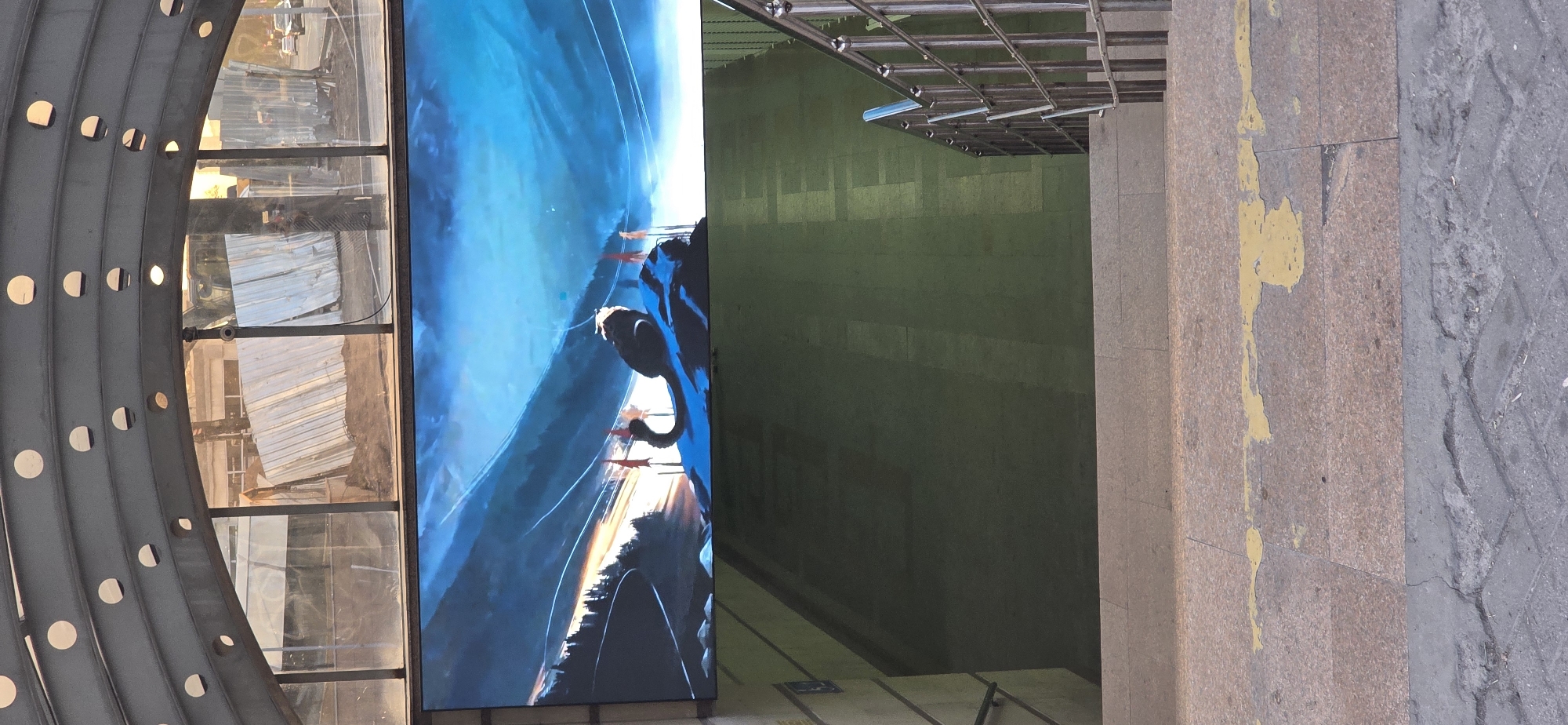 The image shows an outdoor LED screen displaying an animated artwork in a passage or underpass in Bishkek City, Kyrgyzstan. The artwork depicts a fantasy landscape with mountains, trees, and a stylized creature. In the background, through a transparent roof structure, construction is visible with metal sheeting and possibly heavy machinery. The passage itself appears to be an entrance to a subterranean area, with concrete stairs and a metal railing leading downwards. The lighting suggests it is daytime, with sunlight filtering through the transparent roof. The overall scene conveys a sense of urban infrastructure combined with digital art and ongoing development.