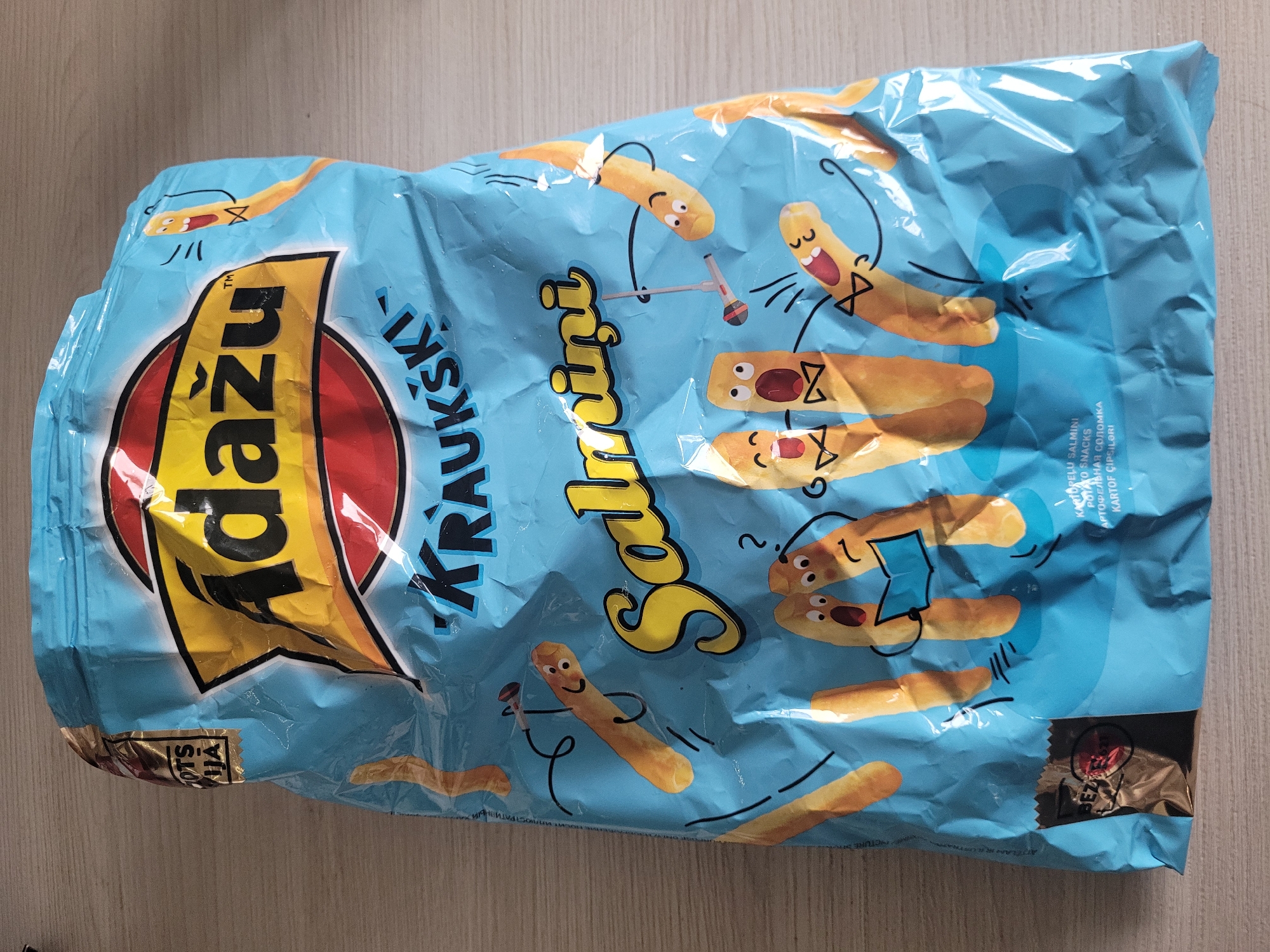 The image shows a bag of "Adazu Kraukšķi Salmini" potato snacks, which is a product from Latvia. The bag is light blue with yellow and red branding. It features cartoonish drawings of potato stick characters, some with mouths open as if singing or shouting. The snacks themselves appear to be thin, elongated, and golden brown. The bag is lying flat on a wooden surface, possibly a table or floor, with a slight sheen indicating the material of the bag. There are some creases and wrinkles on the bag, suggesting it has been handled or is not perfectly taut. In the background, the surface appears to be a light-colored wood grain. The product is a common snack item, not specific to any particular location within Jūrmala, Latvia, beyond being a product of Latvian origin.