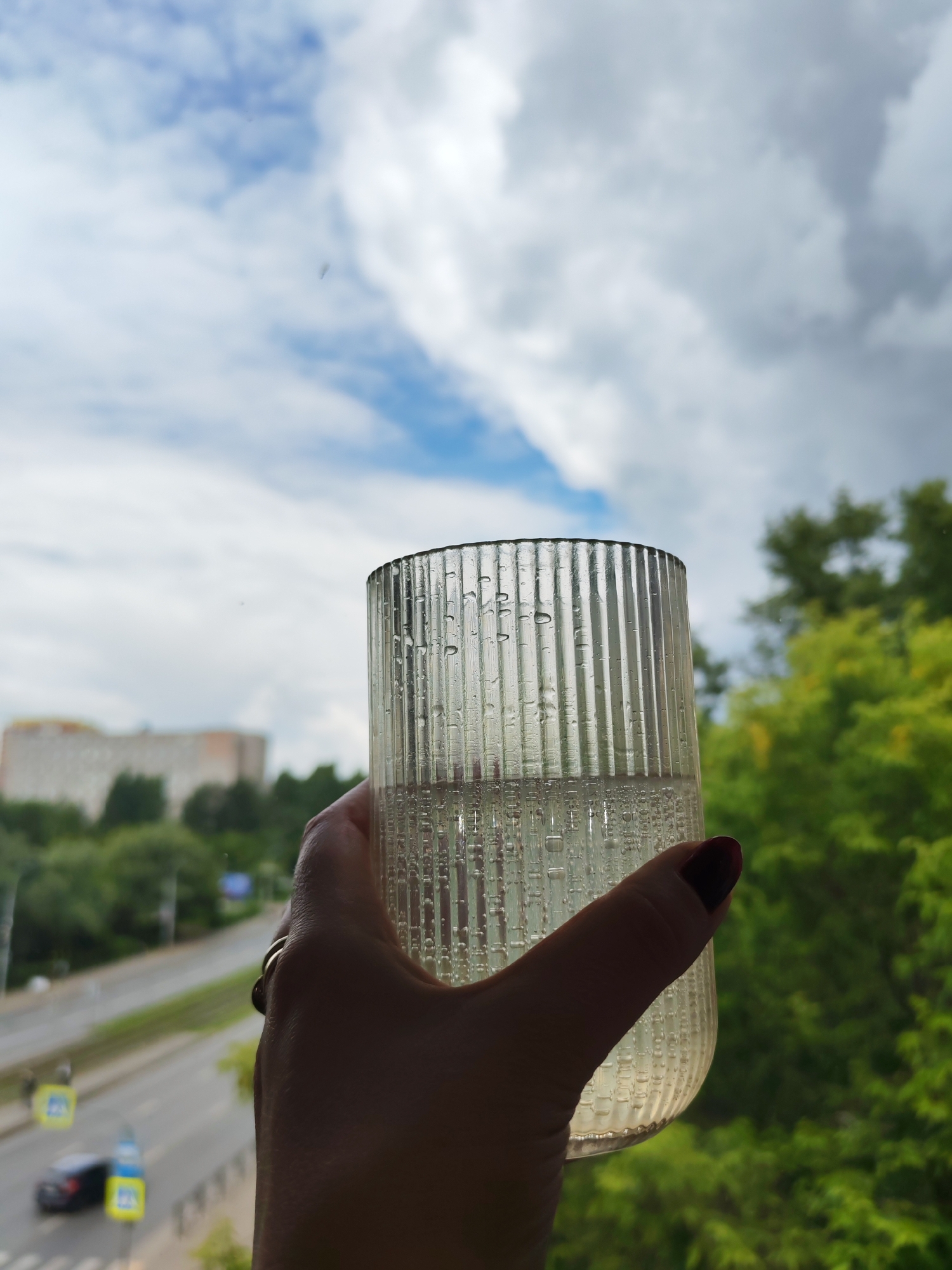 A hand holds a textured glass containing a clear liquid, possibly water, against a backdrop of a cloudy sky and city scenery. The glass has condensation droplets on its exterior, suggesting a cool beverage. In the middle ground, a blurry view of a street with cars and buildings, including a tall apartment complex, is visible. The foreground is dominated by the hand and the glass, obscuring much of the person. The overall lighting suggests it is daytime, with the clouds creating diffused light. The presence of trees in the background indicates a green, possibly park-like area within the urban environment. The style of the buildings and road infrastructure are consistent with urban planning in many Russian cities, including Izhevsk.
