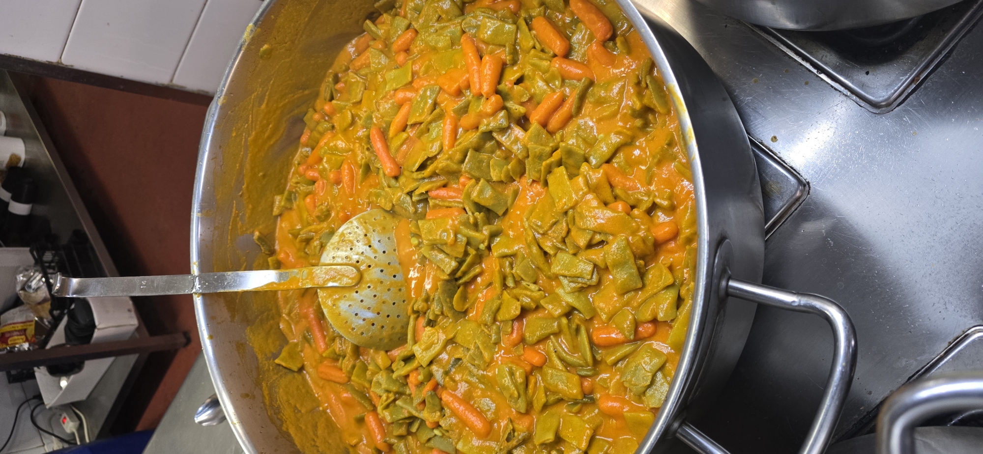 This media file captures a close-up, overhead view of a large pot filled with a vibrant, orange-hued stew containing green, flat beans and small, whole carrots. A metal ladle with numerous perforations rests partially submerged in the stew, suggesting it is being served or prepared. The pot itself is made of stainless steel and appears to be in a professional kitchen environment, indicated by the surrounding stainless steel surfaces and tiled walls.
The lighting suggests it could be daytime, given the brightness of the scene, though no direct sunlight or specific time of day cues are evident. The setting is likely a commercial kitchen or catering establishment.
Visible text is limited to a small "14" stamped on the metal handle of the ladle. There are no people visible in the frame. The overall impression is one of a hearty, freshly prepared dish ready to be served. The context provided, "Sallent de Gállego, Spain," places this scene in a specific geographic location, hinting at local cuisine.