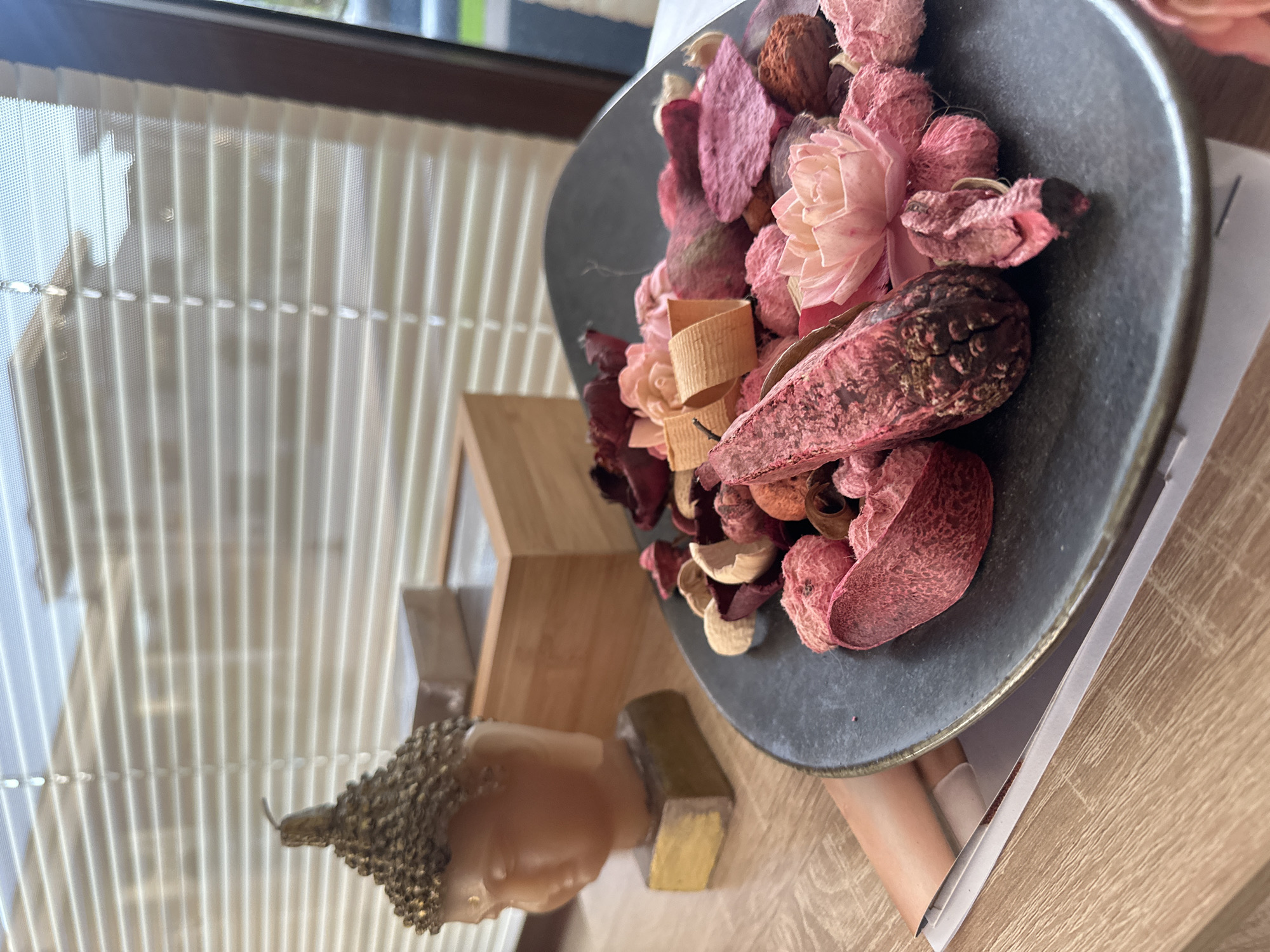 A still life arrangement is depicted on a wooden surface, likely a table, near a window with horizontal blinds. In the foreground, a dark, irregularly shaped plate is filled with a variety of dried botanicals, predominantly in shades of pink, red, and beige. These include what appear to be dried flower petals, seed pods, and possibly small wooden decorative elements. To the left of the plate, a small, stylized bust of Buddha, cast in a golden or brass-like material, sits on a golden base. Behind the Buddha bust, a light-colored wooden box is visible, its lid slightly ajar. The background is dominated by the window and its blinds, through which diffused light enters, suggesting an indoor setting. The scene has a calming, decorative aesthetic, possibly intended for a spa or home decor. The location is identified as Sozopol, Bulgaria, though the image itself does not contain any specific visual cues to confirm this.